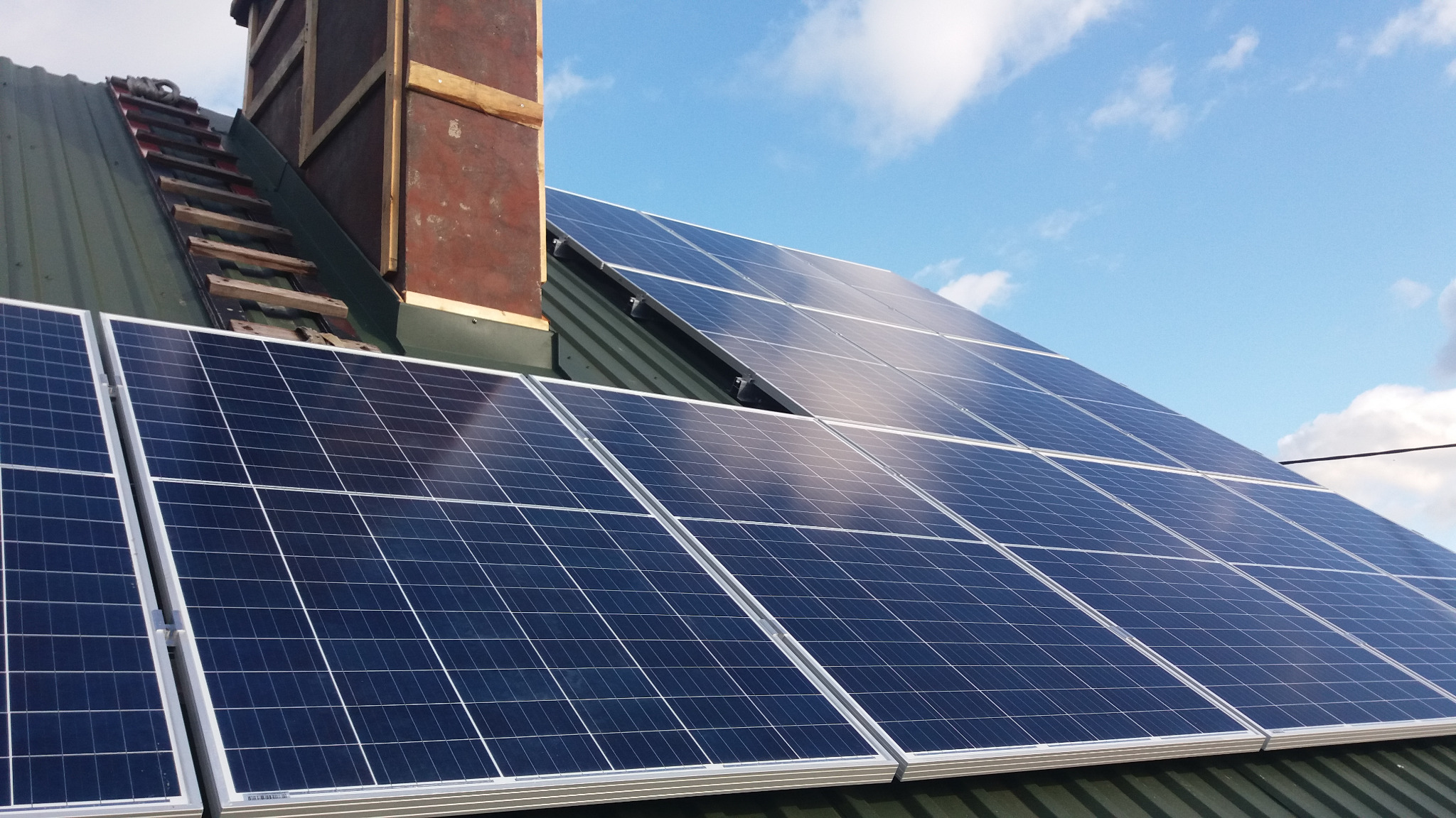 Panele fotowoltaiczne zamontowane na zielonym dachu blisko komina z drewnianą konstrukcją wsporczą, widoczne fragmenty drabiny dachowej i błękitne niebo z chmurami.