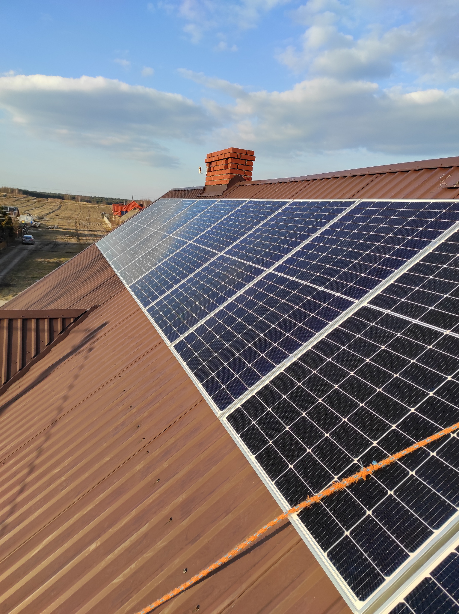 Panele słoneczne na brązowym blaszanym dachu domu z ceglanym kominem, widoczne pole w tle i fragment pomarańczowej liny zabezpieczającej.
