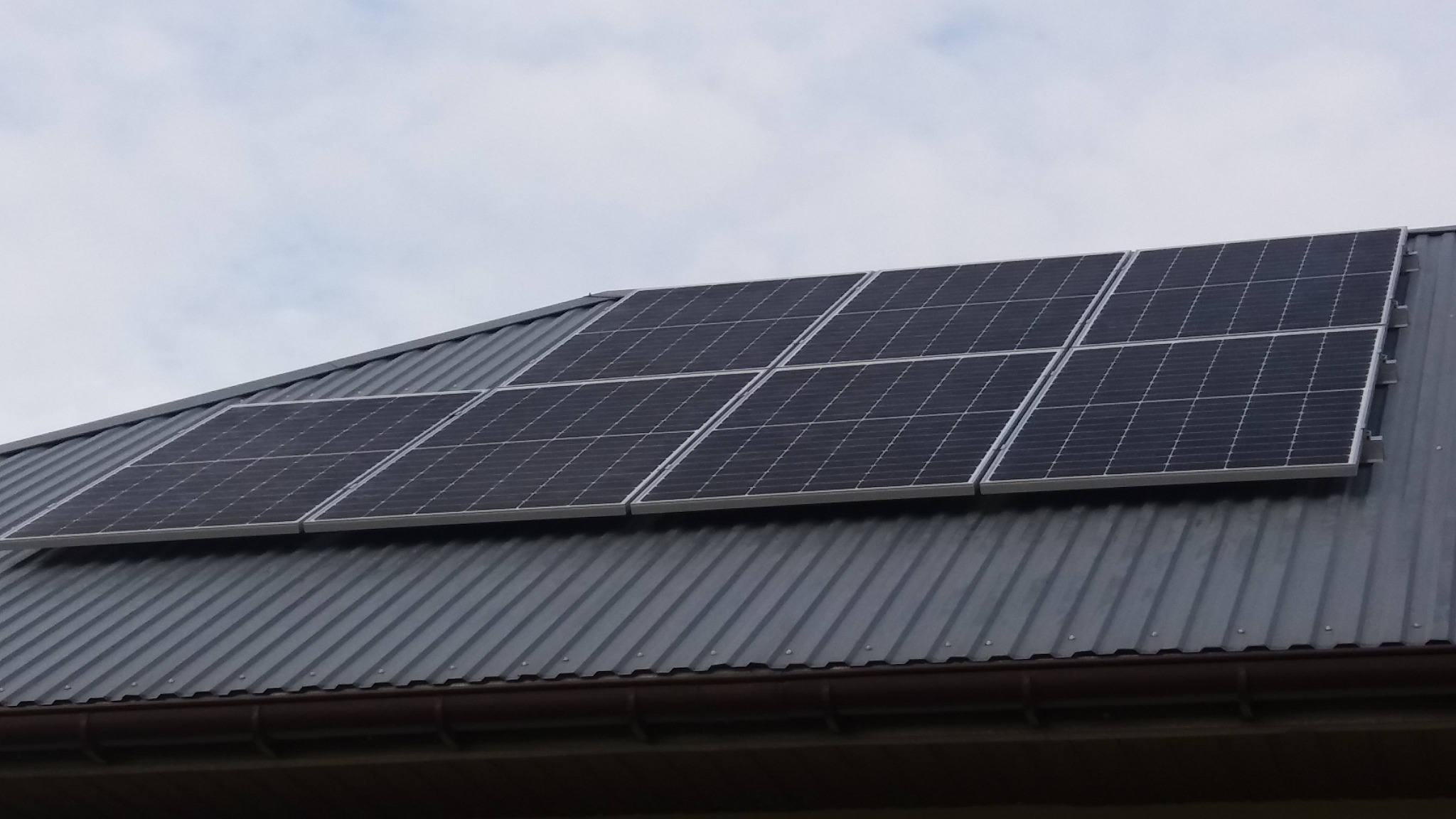 Panele słoneczne na metalowym dachu z rynną, widoczne mocowania paneli, pochmurne niebo w tle.
