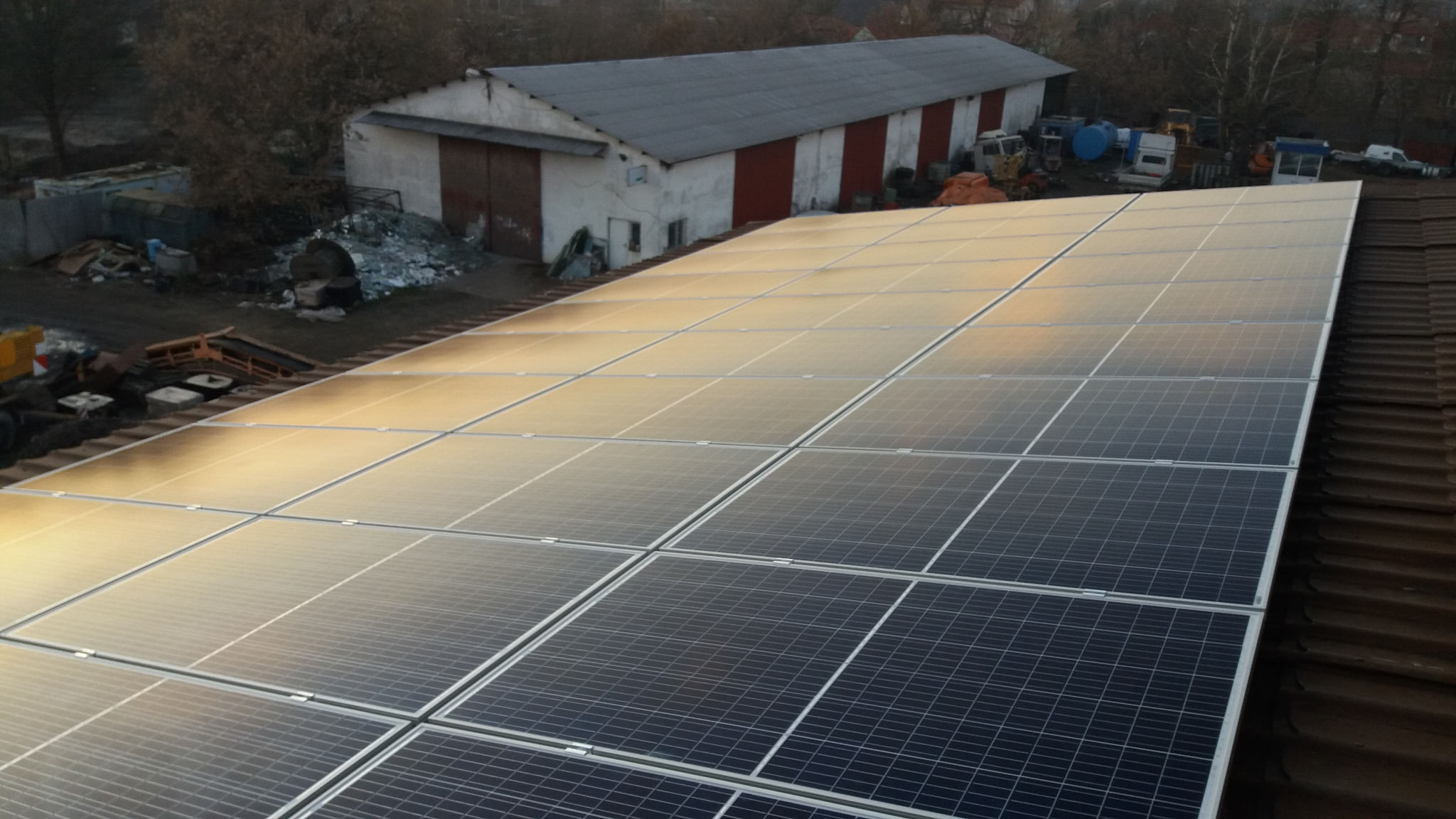 Instalacja paneli słonecznych na dachu budynku gospodarczego z widocznym sprzętem budowlanym w tle, uchwycona z góry w pochmurny dzień.