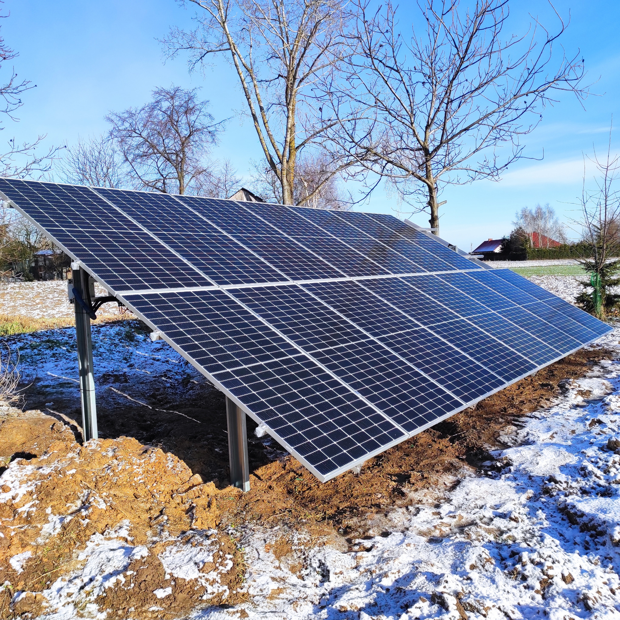 Panel fotowoltaiczny zamontowany na ziemi, z widocznymi śladami po instalacji w postaci rozkopanej ziemi i resztek śniegu, na tle zimowego krajobrazu z drzewami bez liści i domami w oddali.