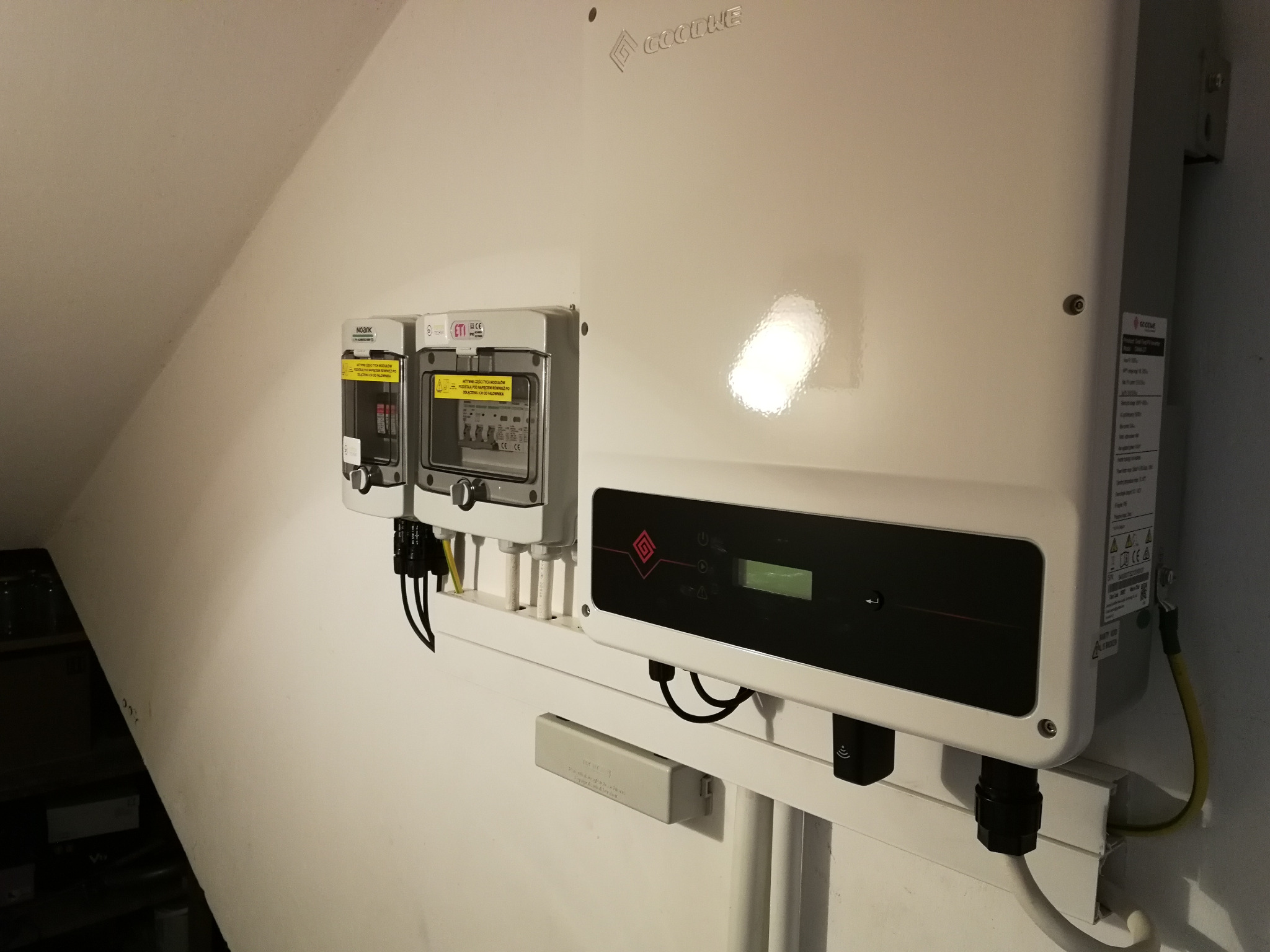 Biały falownik GoodWe z zabezpieczeniami modułowymi, zamontowany na białej ścianie pod skosem strychu, widoczne przewody i elementy montażowe.