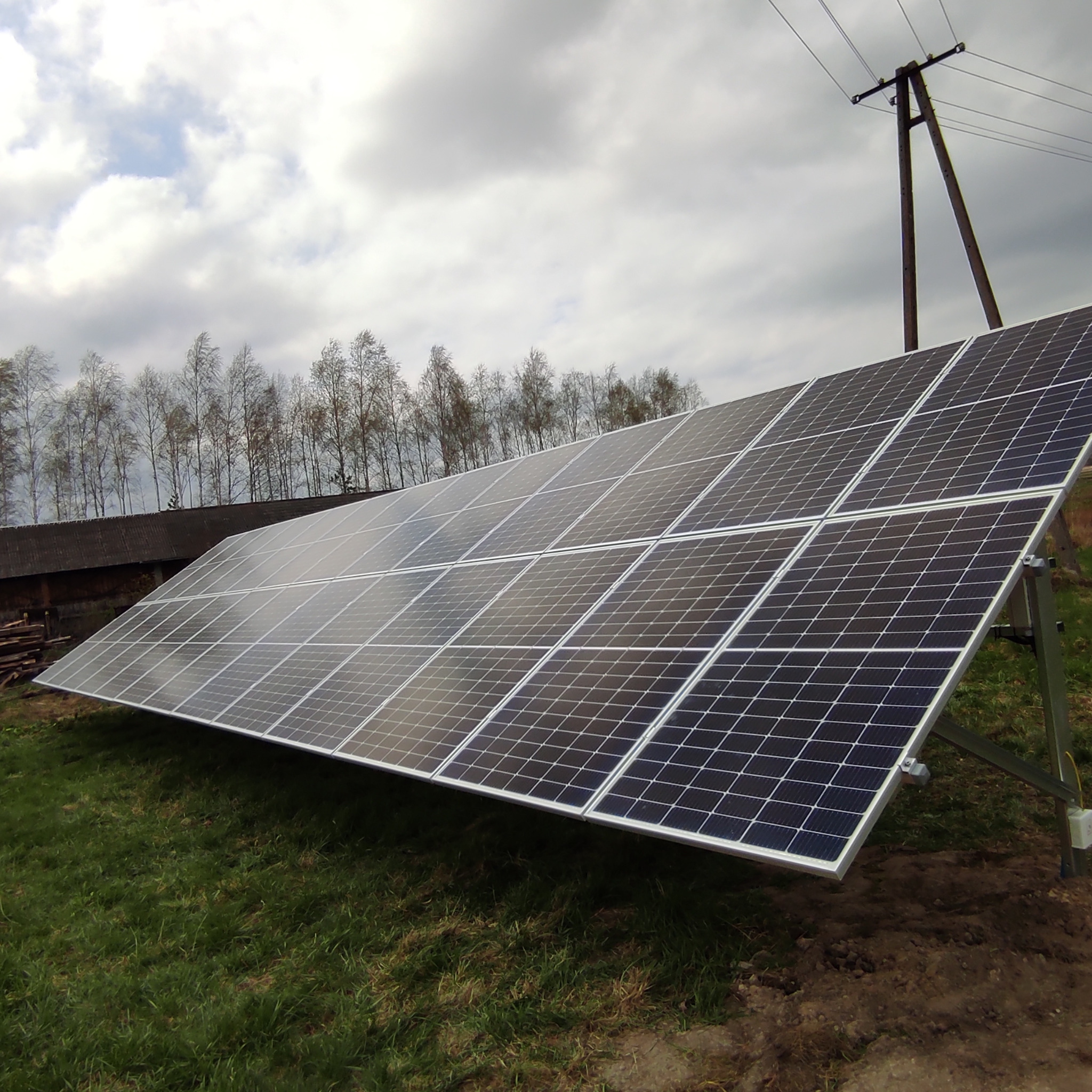 Panel fotowoltaiczny zamontowany na metalowej konstrukcji, na tle budynku gospodarczego i słupa energetycznego, pochmurne niebo.