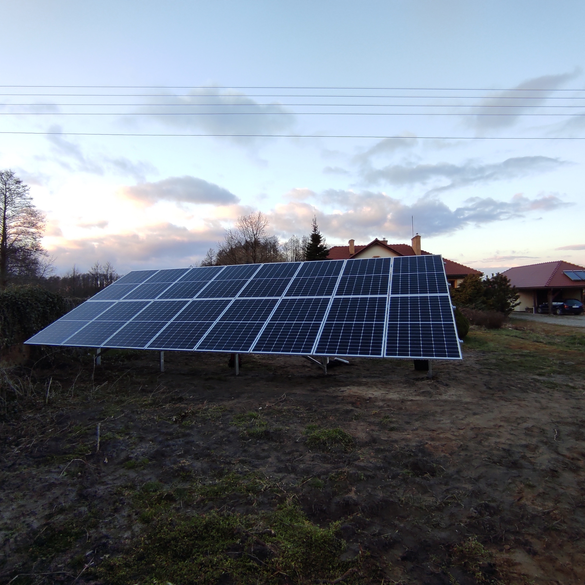 Panel fotowoltaiczny na metalowej konstrukcji, zamontowany na podwórku przed domem jednorodzinnym, widok z dołu na tle zachmurzonego nieba.