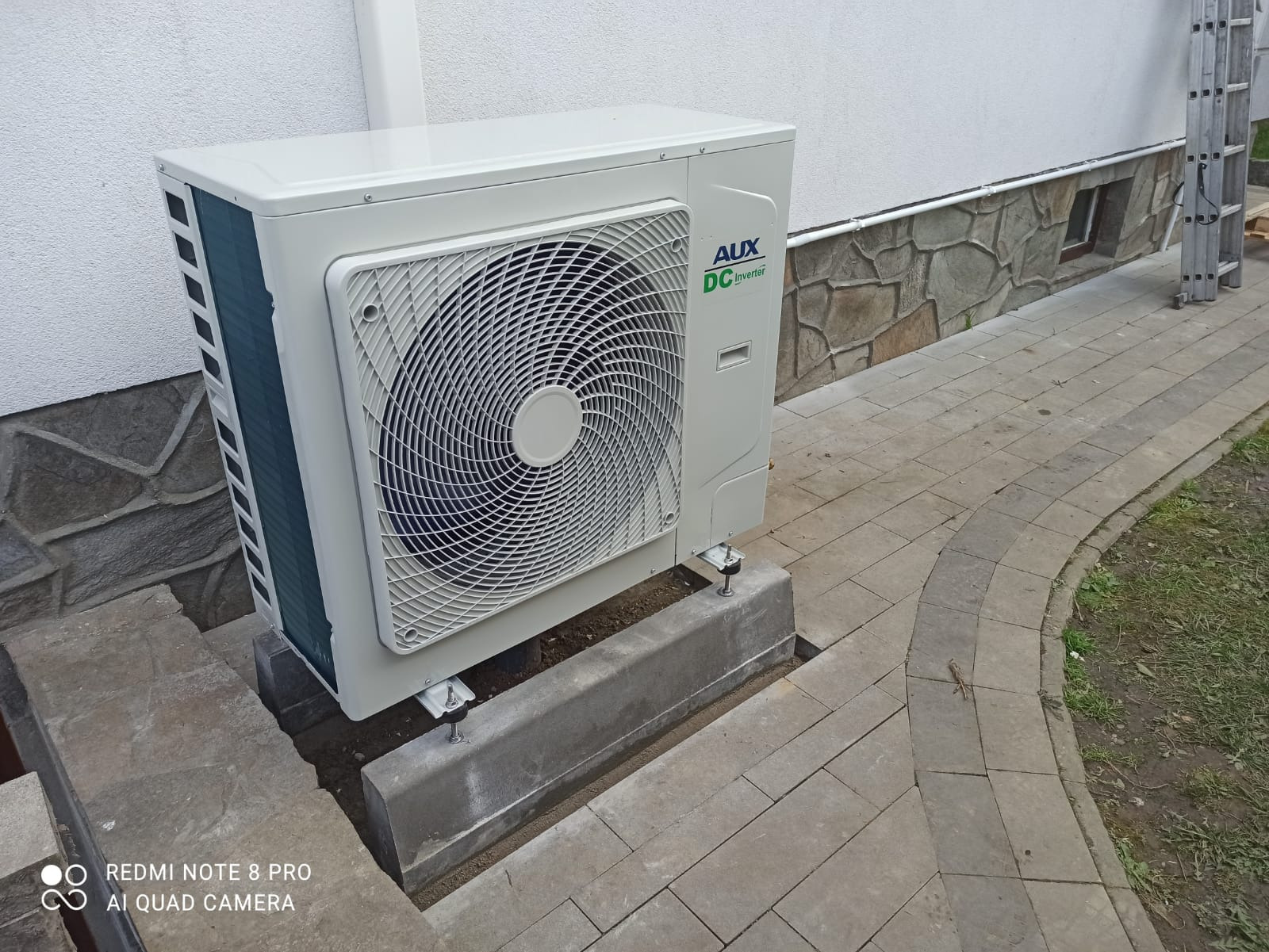 Biała jednostka zewnętrzna klimatyzacji AUX DC Inverter, zamontowana na betonowym fundamencie przy budynku w Nisko. Widoczna drabina oparta o ścianę, kostka brukowa.