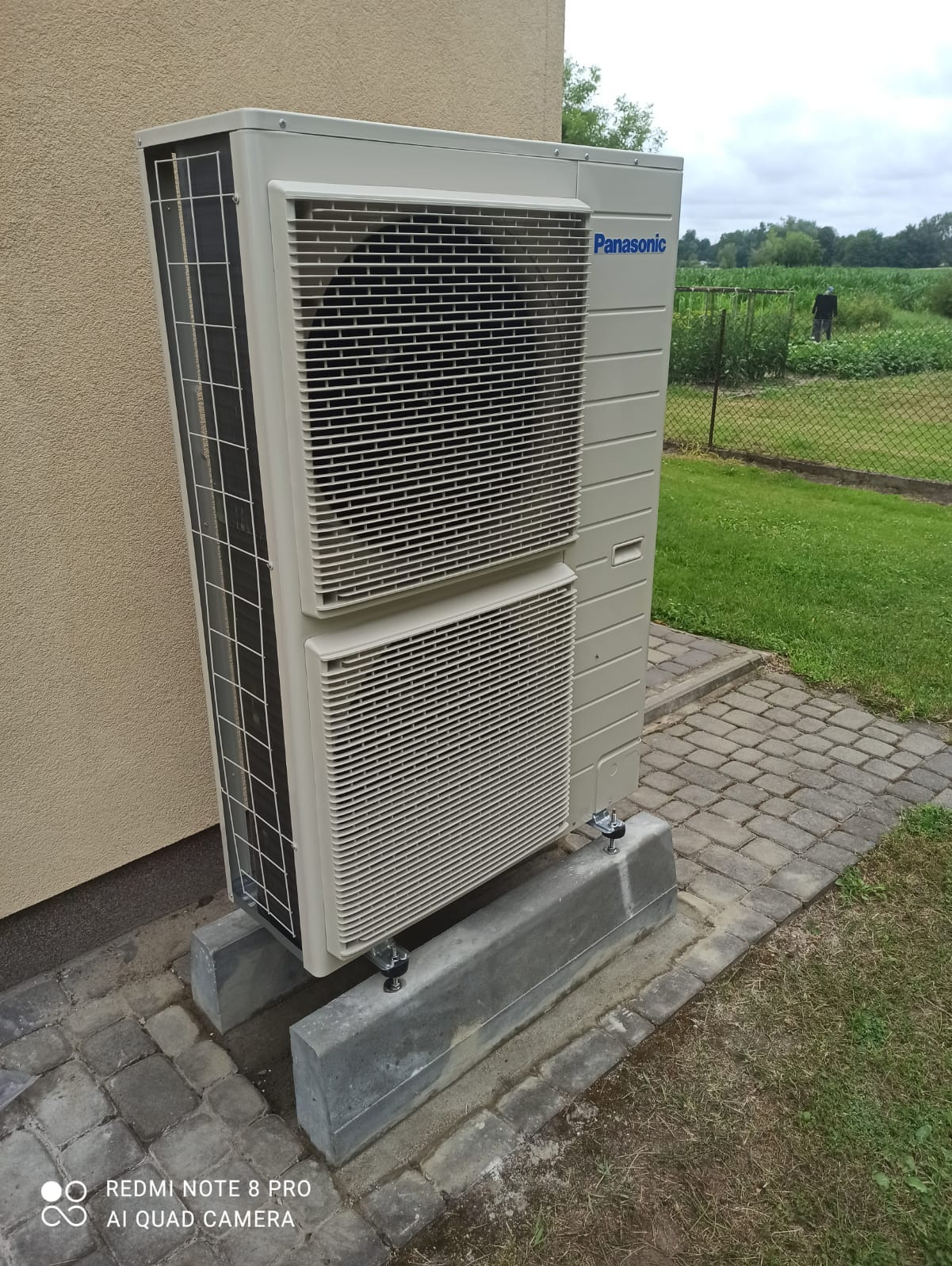 Zewnętrzna jednostka klimatyzacji Panasonic, zamontowana na betonowych wspornikach przy ścianie budynku, z widokiem na zielony ogród w tle.