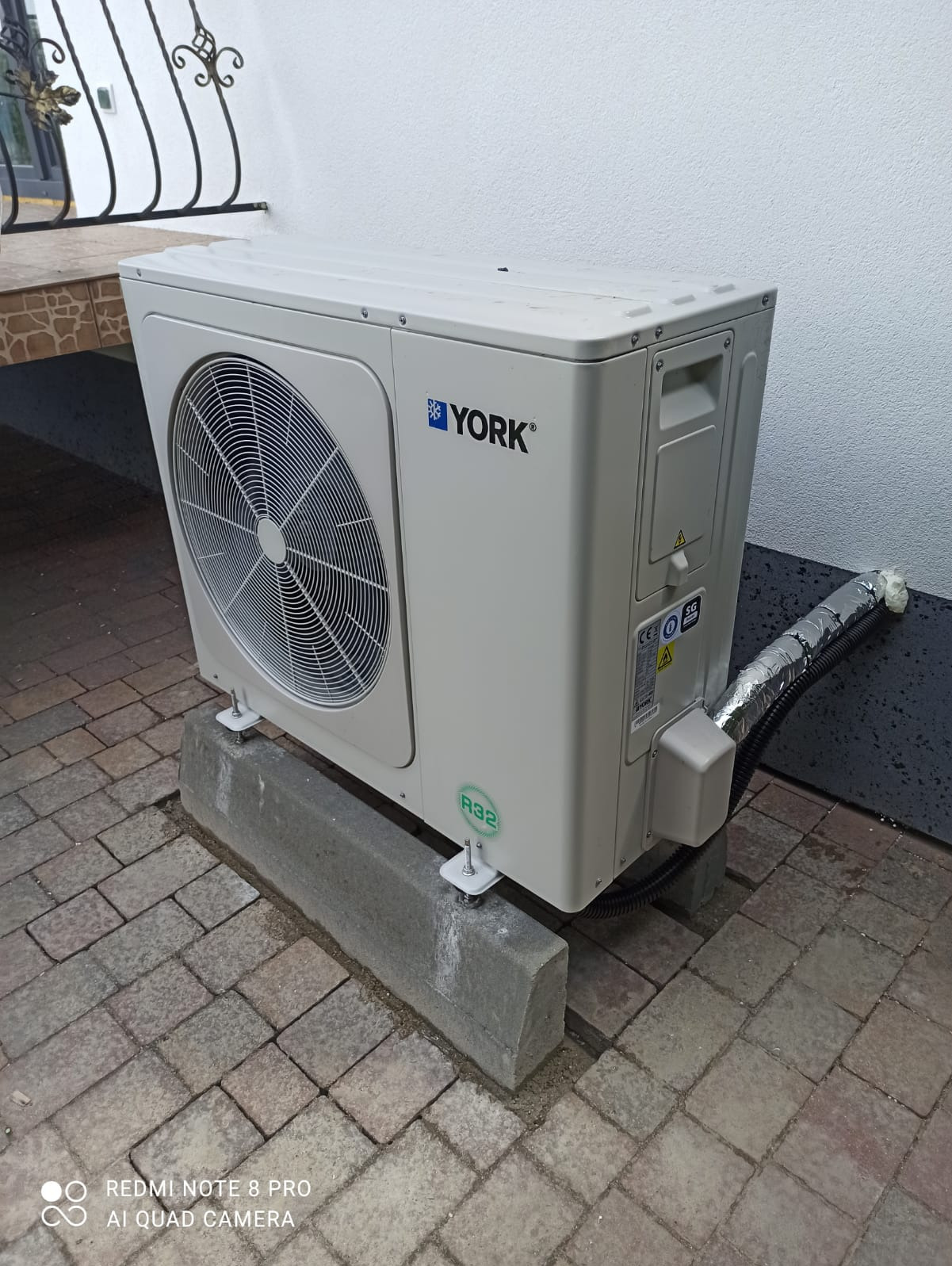 Biała jednostka zewnętrzna klimatyzacji York, umieszczona na betonowych podstawach, z widoczną instalacją i ścianą w tle. Detal montażu.