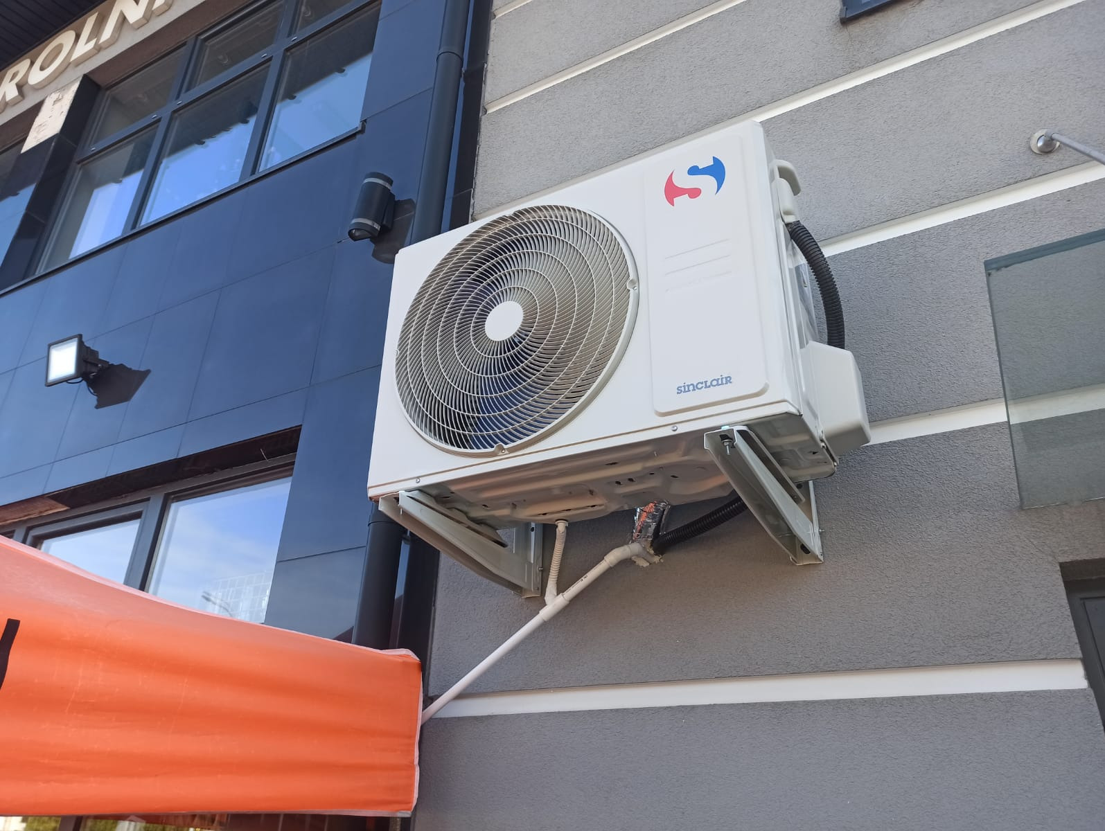 Biały klimatyzator Sinclair zamontowany na szarej elewacji budynku z widocznymi przewodami instalacji. Część pomarańczowej markizy w lewym dolnym rogu kadru.