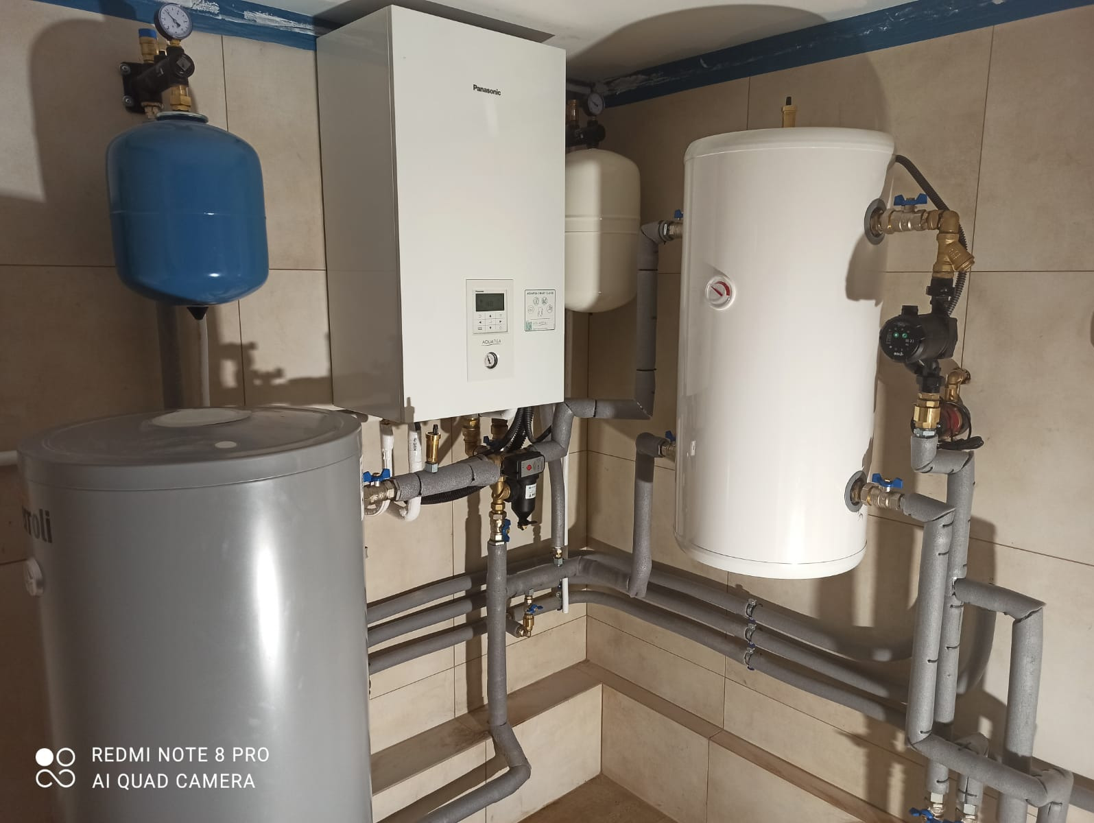 Zestaw urządzeń: biały piec Panasonic, szary zbiornik, biały zasobnik, niebieski zbiornik wyrównawczy, rury w izolacji, manometry. Instalacja w kotłowni.