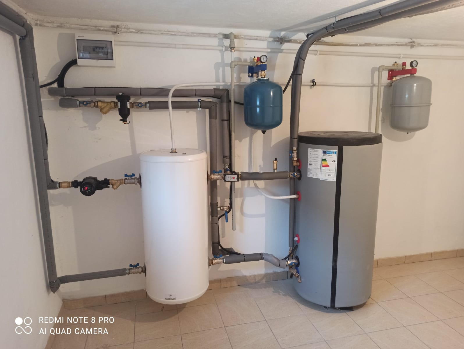 Zestaw zbiorników i rur w kotłowni, widoczne białe i szare zasobniki, instalacja z izolowanymi rurami, manometry i zawory, całość zamontowana na tle białej ściany.