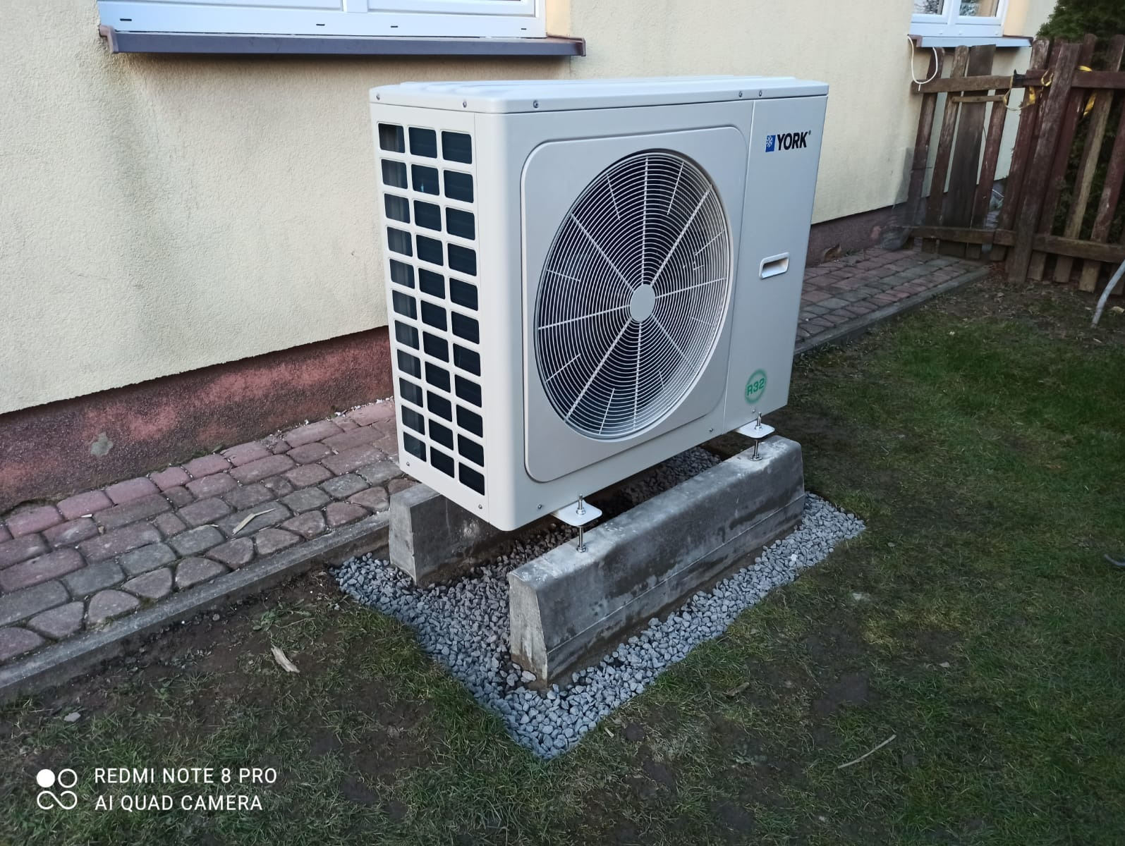 Zewnętrzna jednostka klimatyzacji York, zamontowana na betonowych podstawach, obok budynku z beżową elewacją. Widoczny trawnik i kostka brukowa.