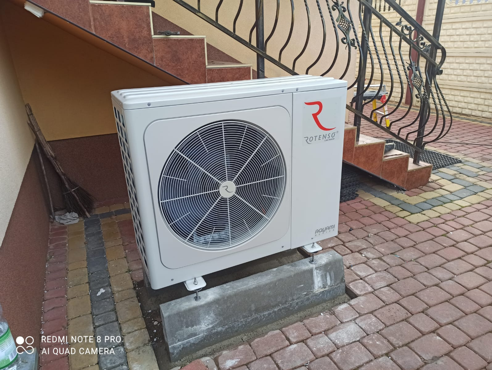 Biała jednostka zewnętrzna klimatyzacji Rotenso Aquami, zamontowana na betonowej podstawie przy schodach z kutą balustradą, na tle kostki brukowej.