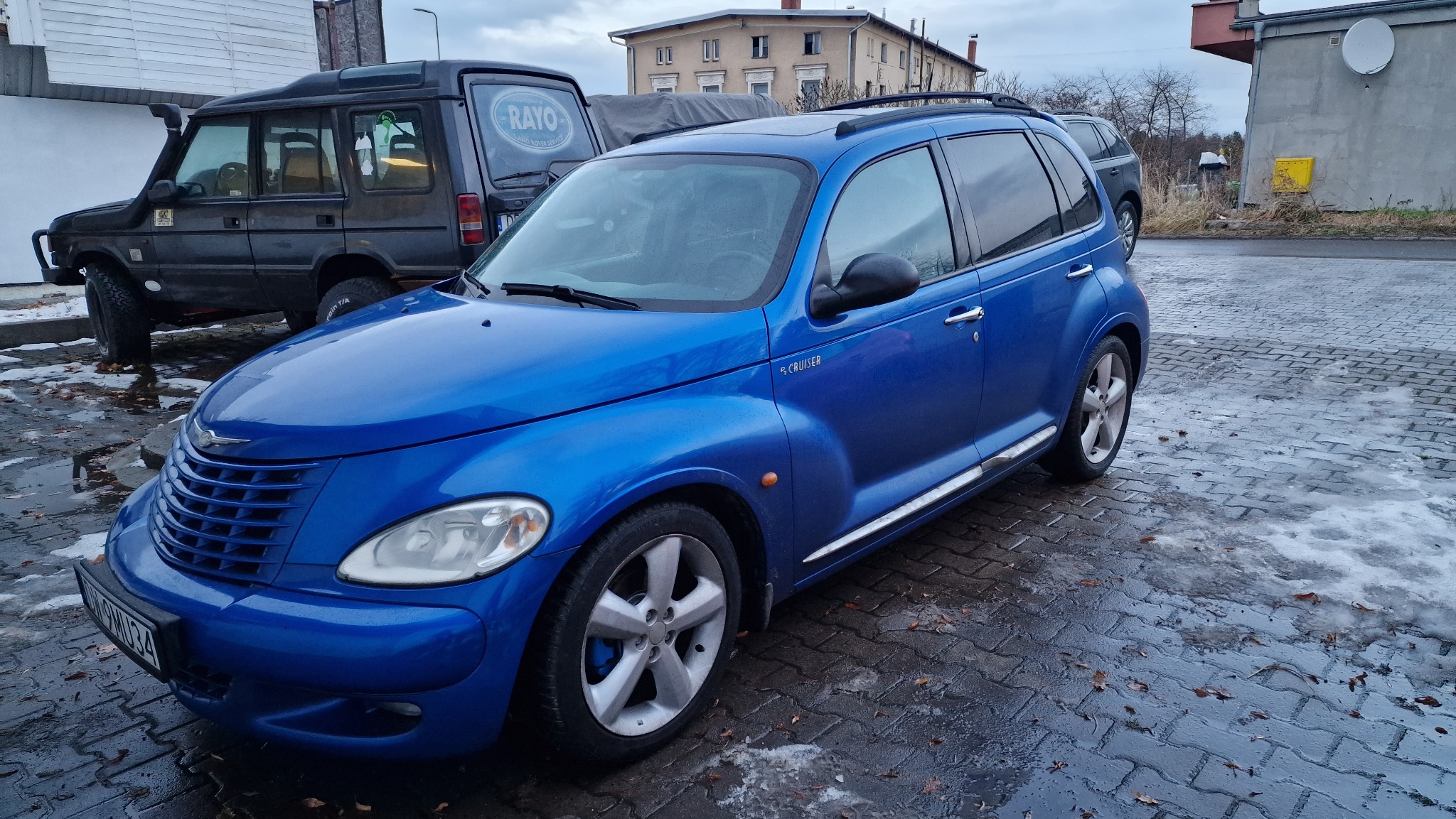 Błękitny Chrysler PT Cruiser zaparkowany na mokrym bruku, widoczny z przodu, z boku i lekko z góry, z innym samochodem w tle.