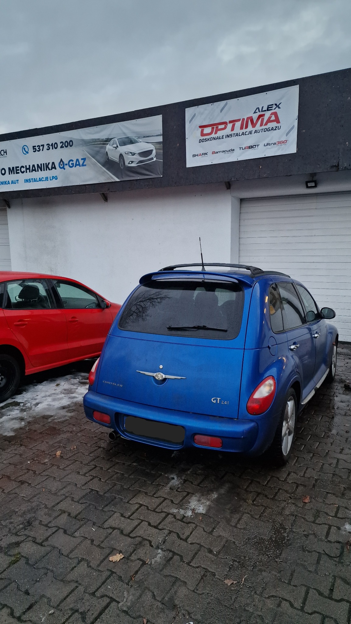 Niebieski Chrysler PT Cruiser GT zaparkowany przed warsztatem samochodowym z reklamą instalacji LPG i mechaniki pojazdowej, widoczny również czerwony Volkswagen Polo.