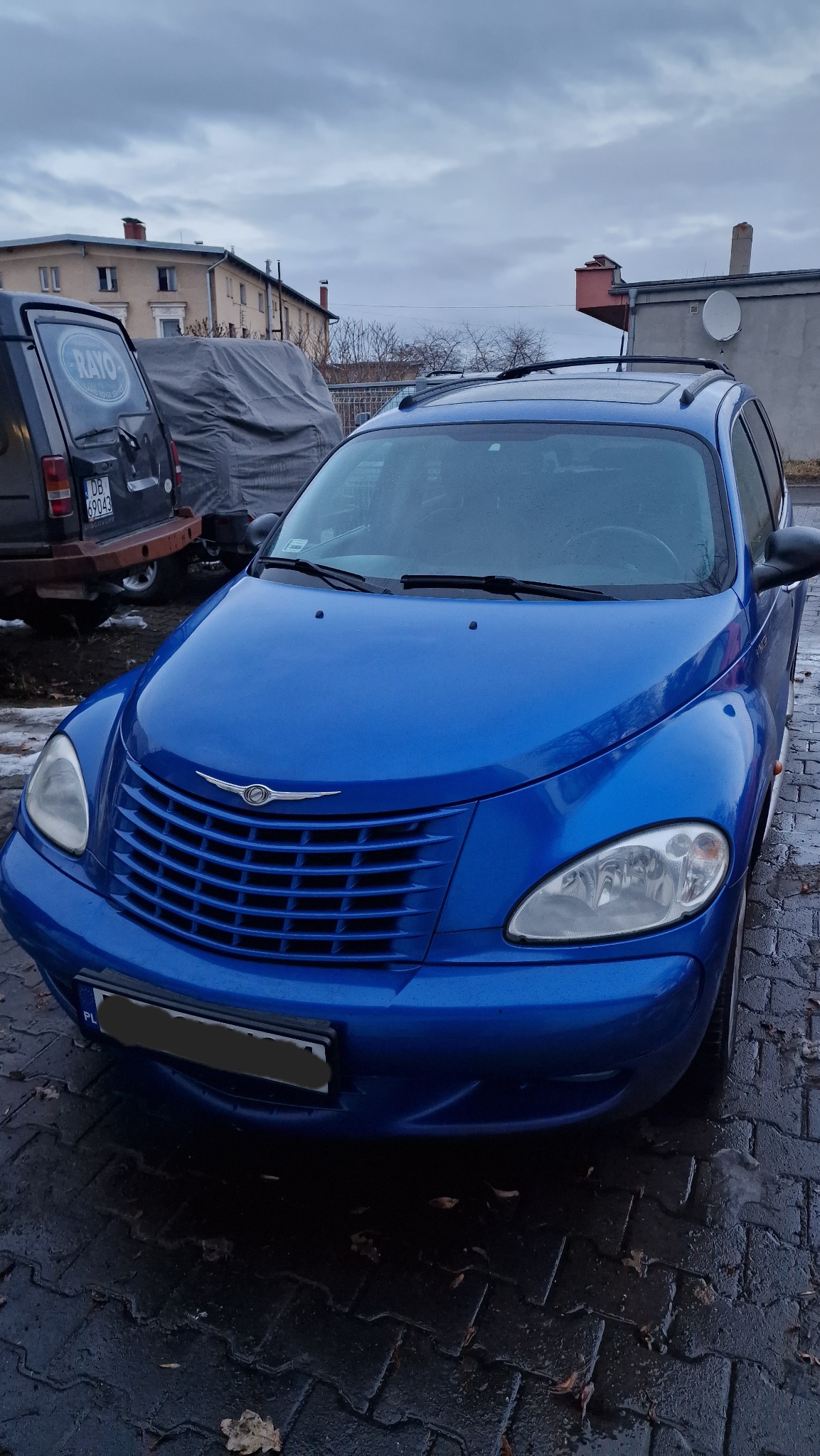 Niebieski Chrysler PT Cruiser na mokrym bruku, widok z przodu, zarys budynków w tle i samochód z logiem RAYO.