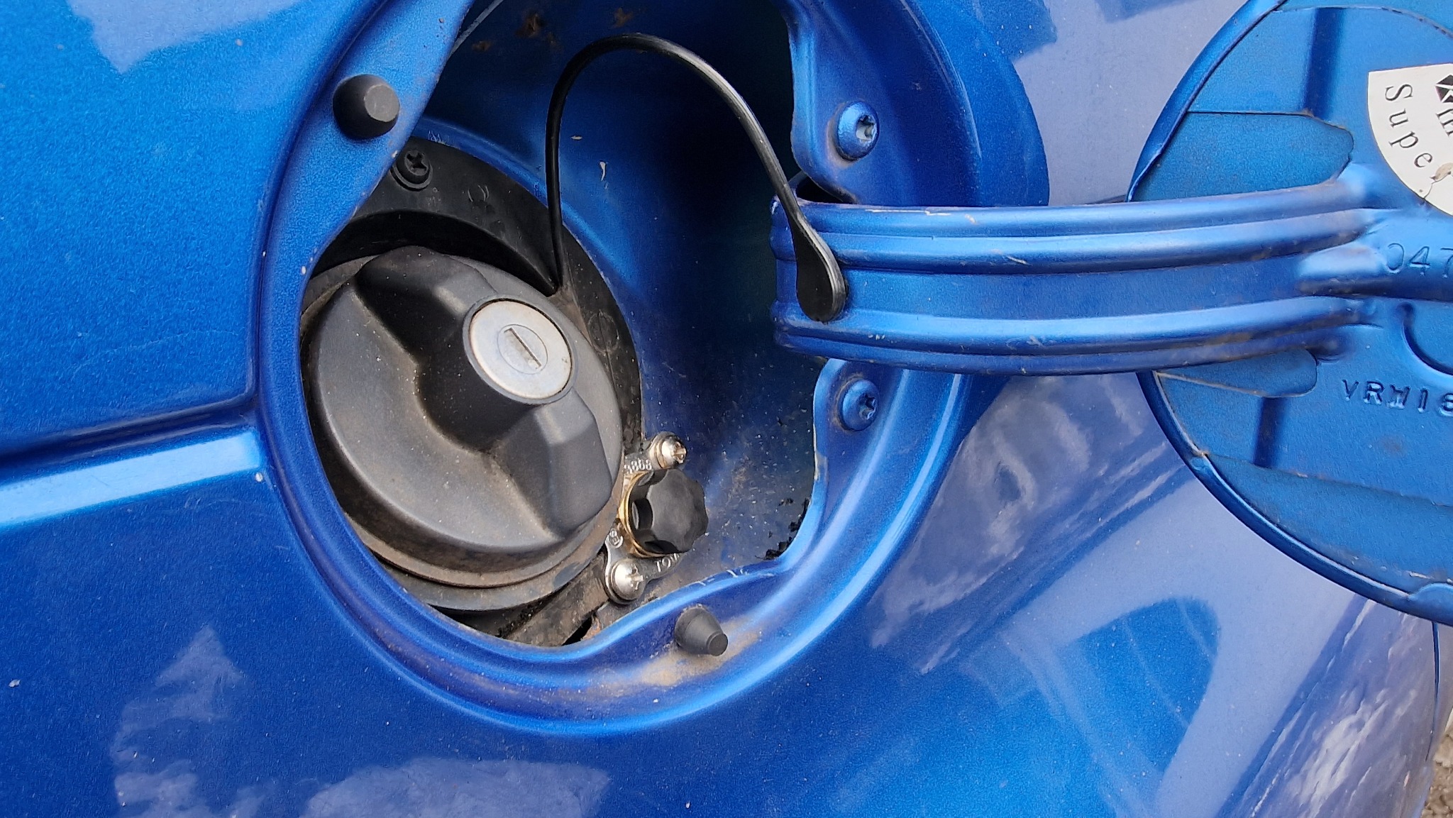 Zbliżenie wlewu paliwa w niebieskim samochodzie z widocznym czarnym korkiem, metalową klapką i naklejką informacyjną.