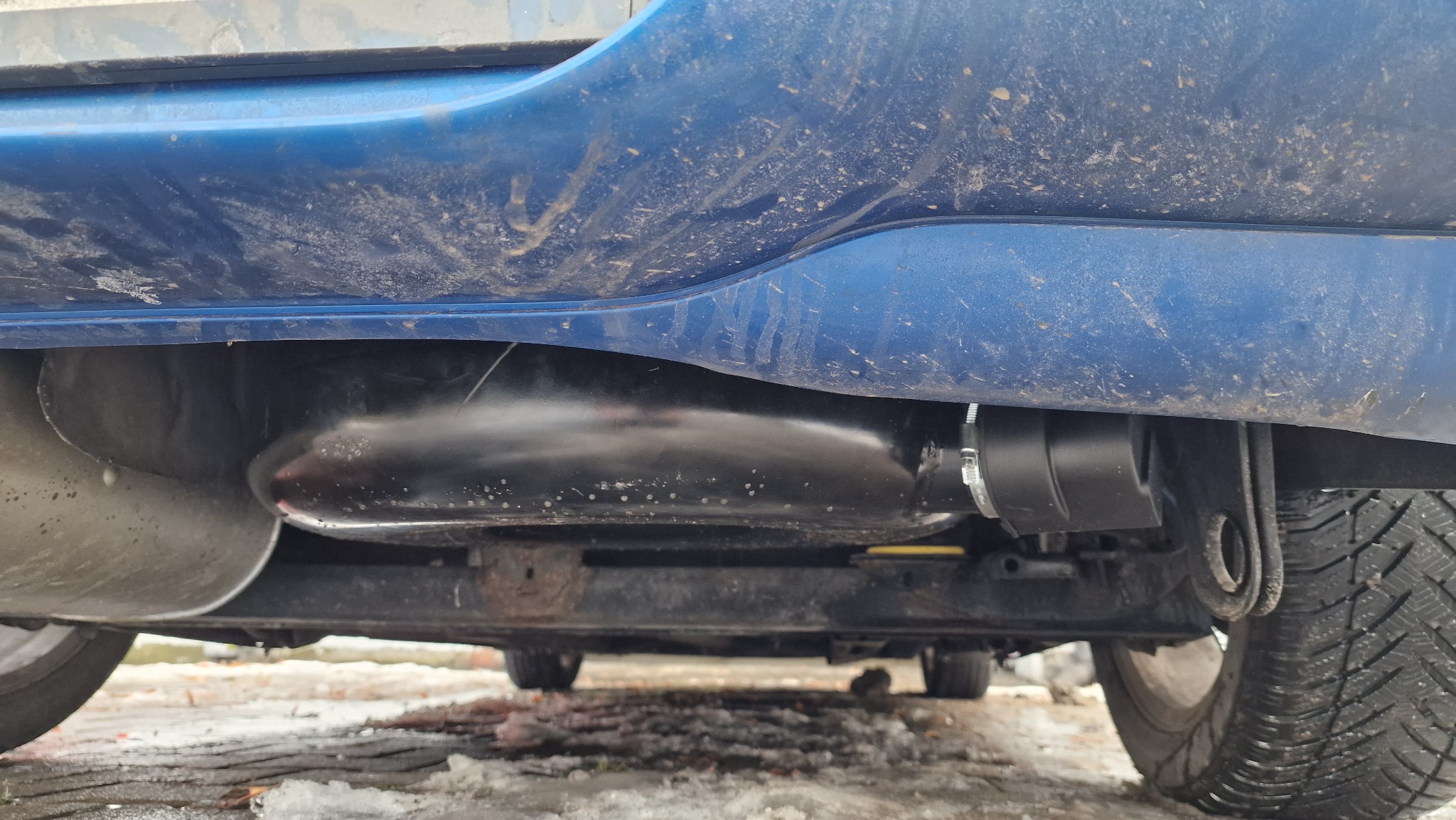 Zamontowany zbiornik LPG pod podwoziem brudnego, niebieskiego samochodu, widoczne opony zimowe i elementy zawieszenia.