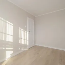 Pusty pokój po remoncie z jasnymi ścianami, drewnianą podłogą i białymi drzwiami, na ścianie widoczne cienie okna.