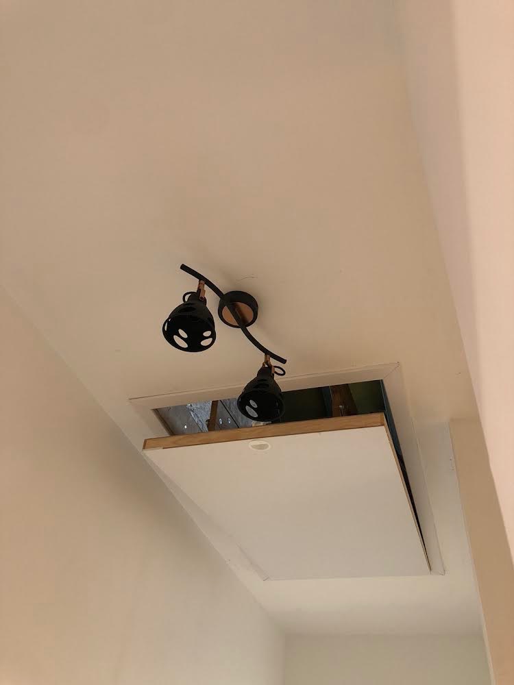 Biała klapa włazu strychowego, uchylona pod kątem, z widocznym wnętrzem strychu, oświetlona czarną lampą sufitową z dwoma reflektorami.