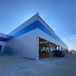 Nowo wybudowany budynek magazynowy z szarymi ścianami i niebieskim pasem, z widocznymi maszynami budowlanymi wewnątrz, niebieski bus zaparkowany z boku, sfotografowany w słoneczny dzień.