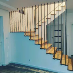 Drewniane schody z metalowymi, pionowymi balustradami w nowoczesnym wnętrzu, widok z dołu, ściana z białej cegły w tle.