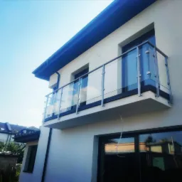 Nowoczesny balkon z przeszkloną balustradą, widok z dołu ukazujący minimalistyczny design i srebrne elementy konstrukcyjne na tle błękitnego nieba.