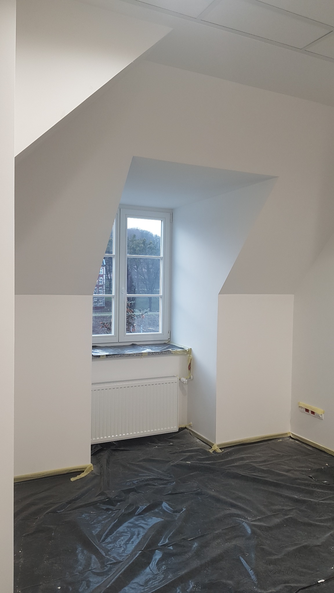Poddasze po odświeżeniu: białe ściany, zabezpieczona podłoga folią malarską, okno z widokiem na drzewa.