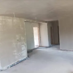 Wnętrze w stanie deweloperskim po nałożeniu gładzi gipsowej, z widocznymi przewodami elektrycznymi wystającymi z sufitu i wężami do ogrzewania podłogowego przy ścianach.