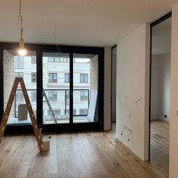 Patryk Wie - Wnętrze mieszkania w trakcie prac wykończeniowych: drewniana podłoga, drabina, widoczne gniazdka elektryczne bez osprzętu, surowa żarówka na kablu, okno balkonowe z widokiem na budynek.