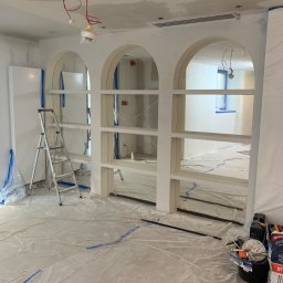 Patryk Wie - Wnętrze w trakcie prac wykończeniowych: ściany po gładzi, zabudowa z półkami w kształcie łuków, podłoga zabezpieczona folią, drabina i narzędzia malarskie.