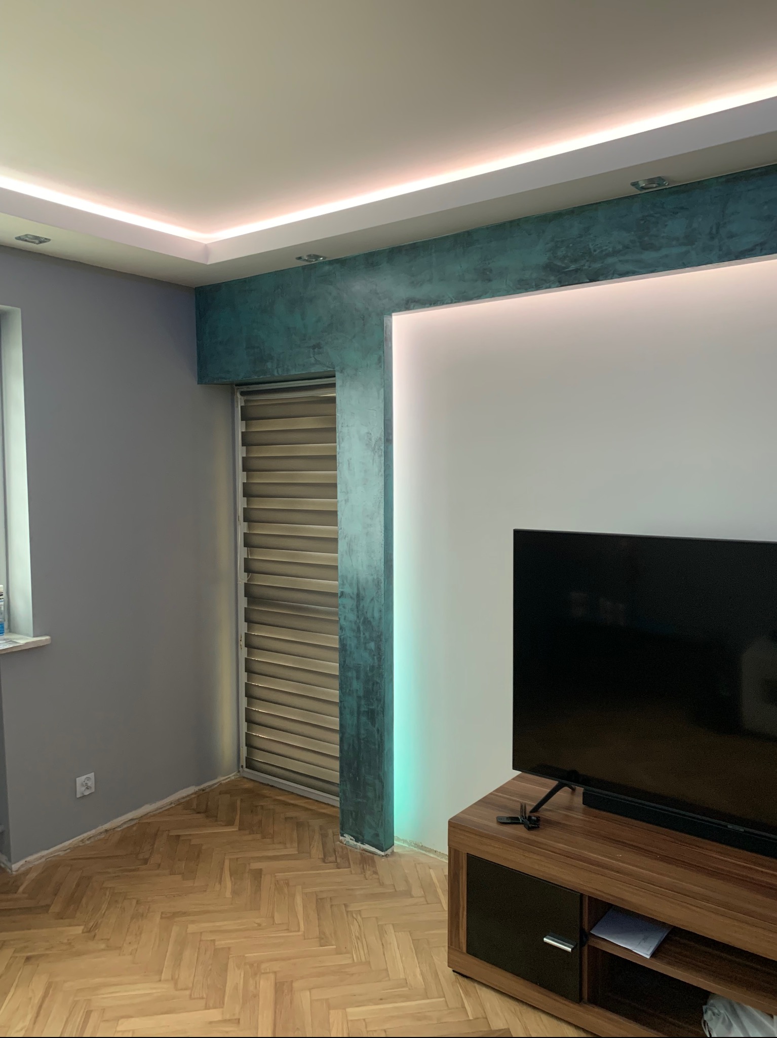 Nowoczesne wnętrze z parkietem w jodełkę, ścianą w odcieniach szarości i turkusu, podświetlanym sufitem oraz telewizorem na drewnianej szafce RTV.