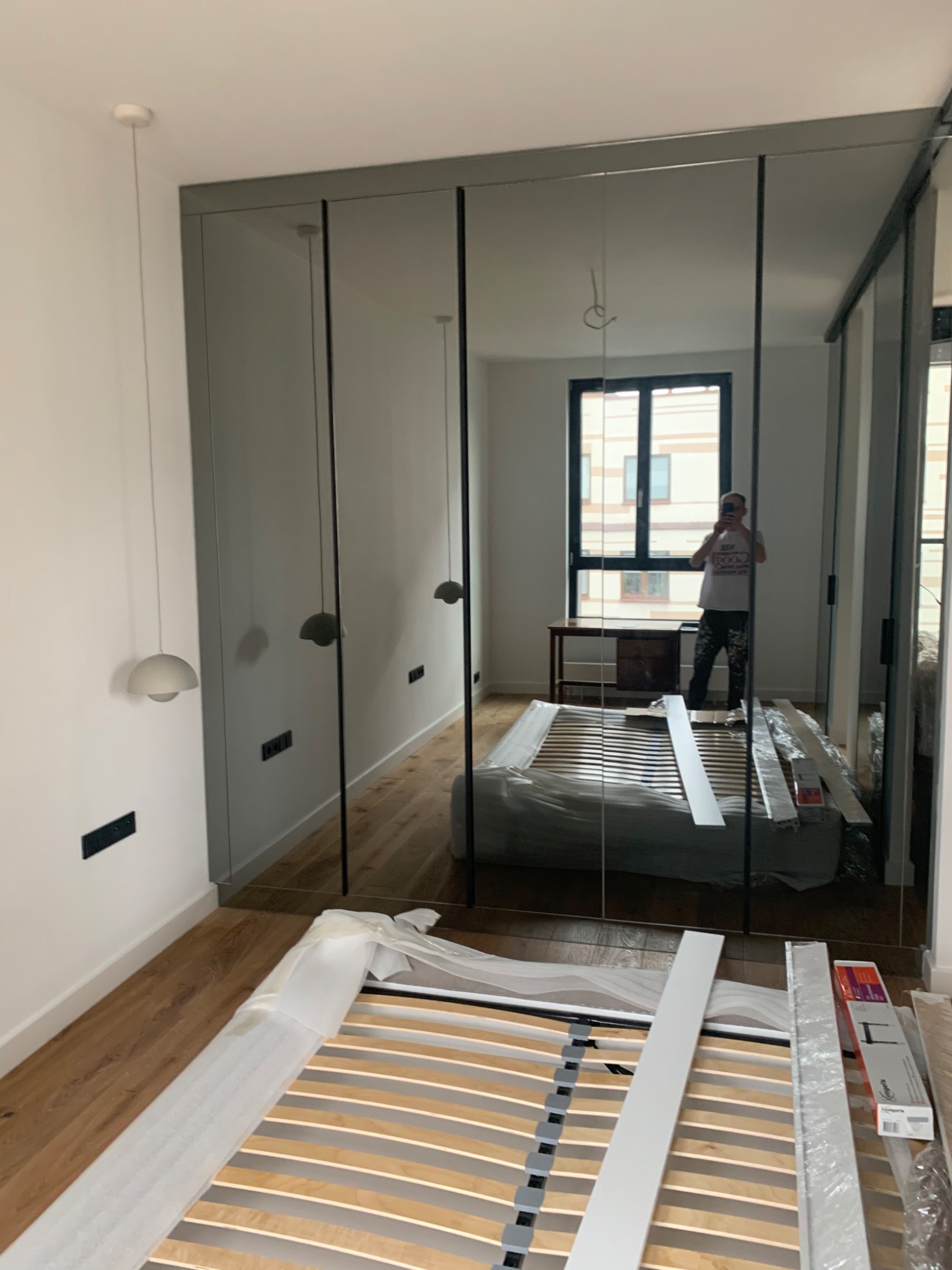 Montaż lustrzanych drzwi przesuwnych do szafy wnękowej w sypialni, widoczny stelaż łóżka w folii ochronnej i osoba robiąca zdjęcie w odbiciu.