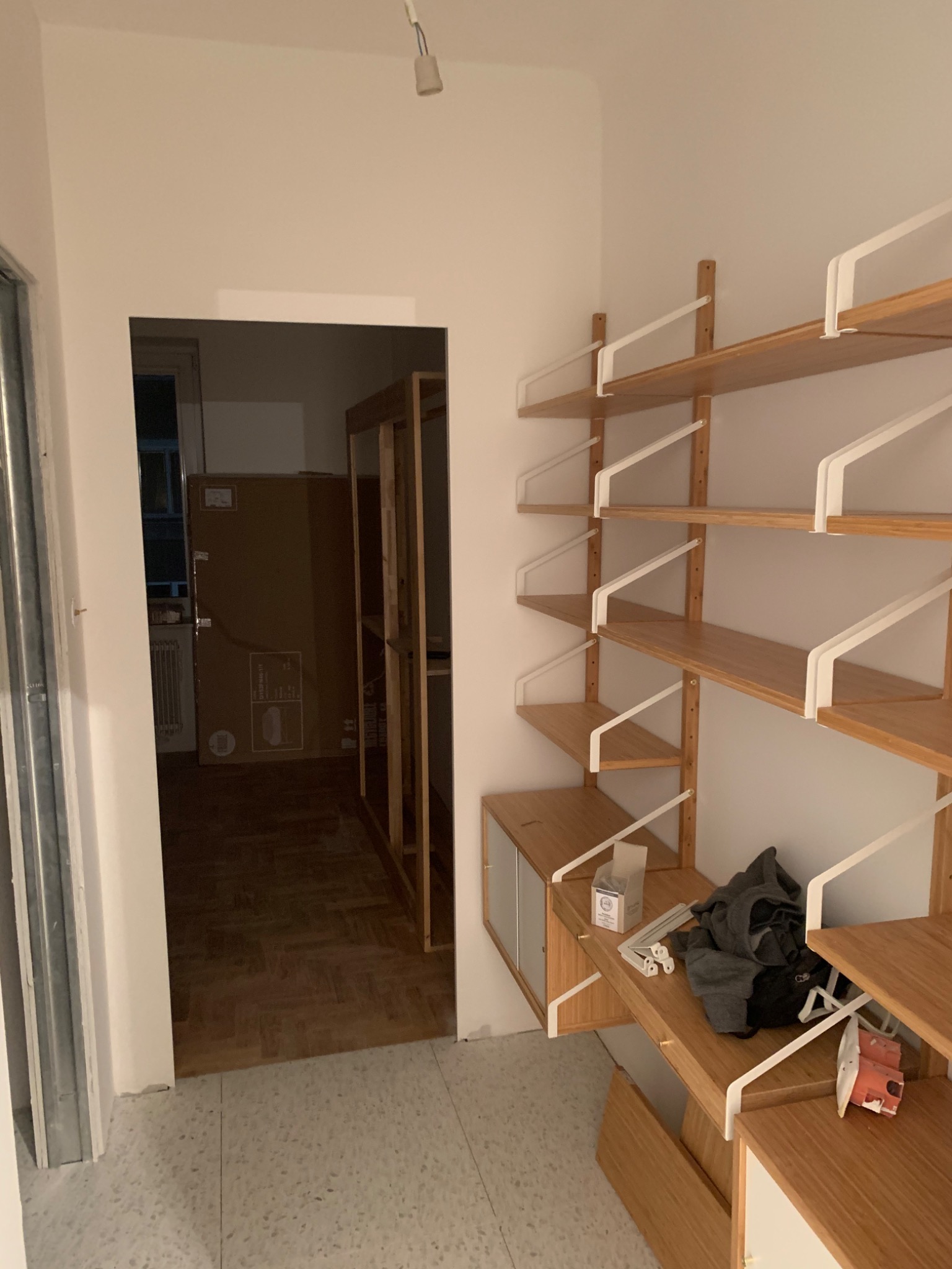 Montaż drewnianego regału modułowego z białymi wspornikami w trakcie prac wykończeniowych mieszkania, widoczne narzędzia i elementy montażowe na półkach, w tle otwarte drzwi do innego pomieszczenia...