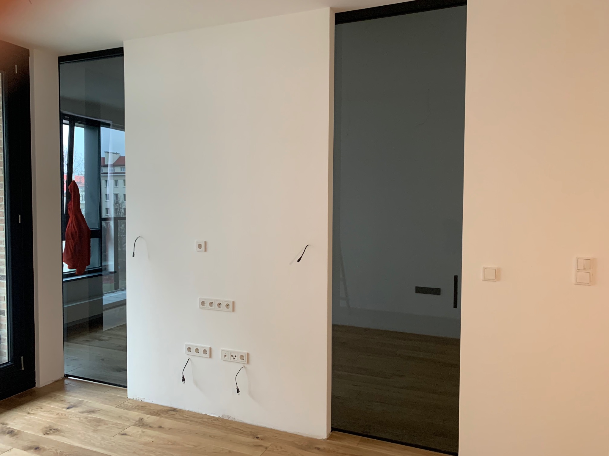Wnętrze mieszkania w trakcie prac wykończeniowych: widoczne gniazdka elektryczne wystające ze ściany, świeżo położona drewniana podłoga oraz przesuwne, szklane drzwi w czarnej ramie.