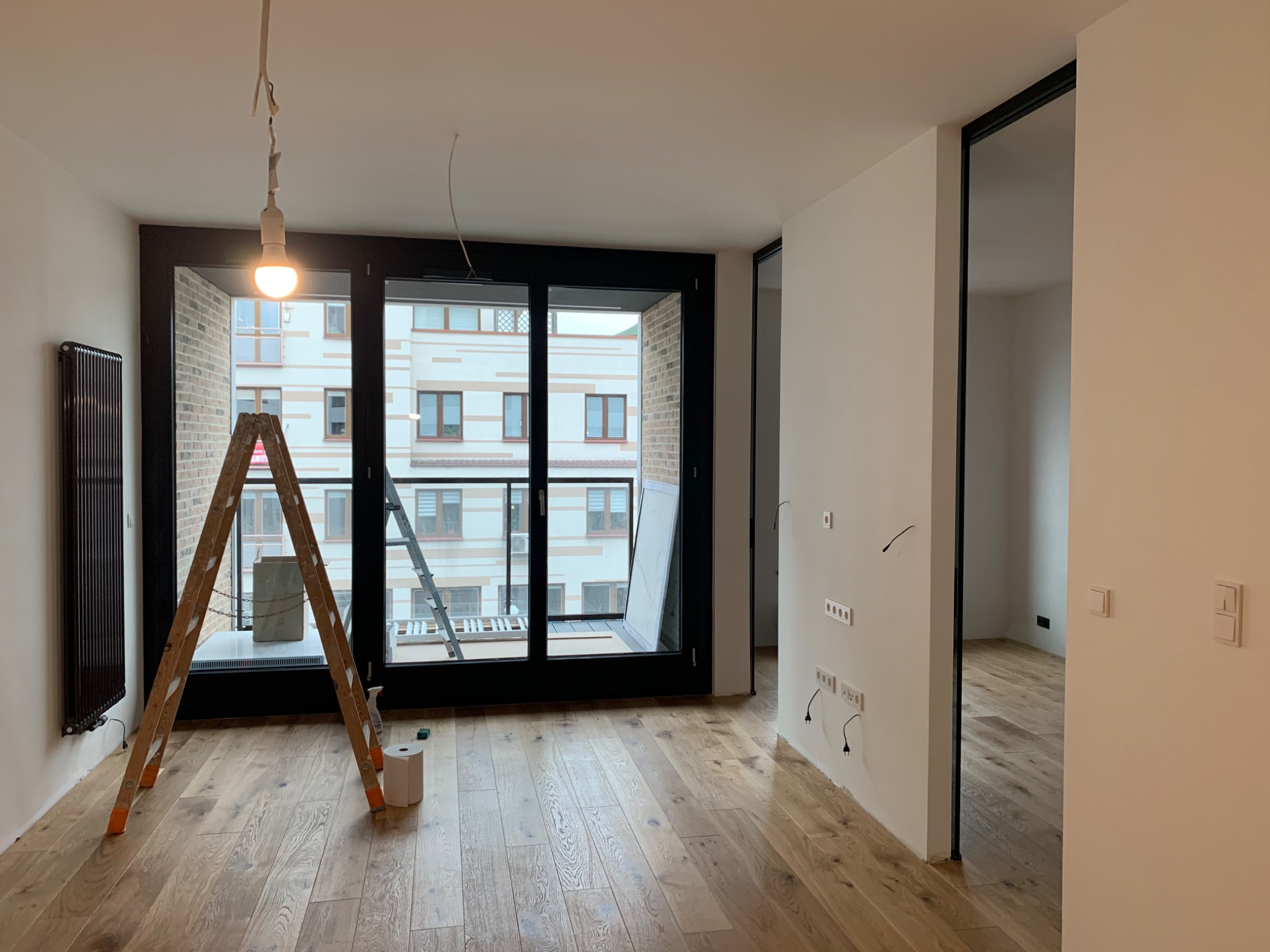Wnętrze mieszkania w trakcie prac wykończeniowych: drewniana podłoga, drabina, widoczne gniazdka elektryczne bez osprzętu, surowa żarówka na kablu, okno balkonowe z widokiem na budynek.