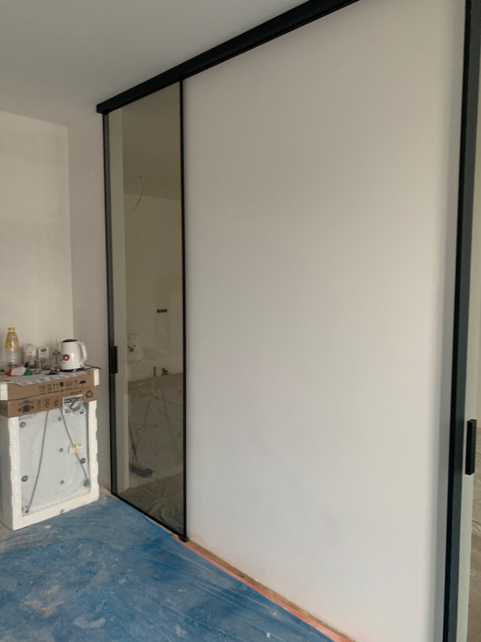 Montaż przesuwnych drzwi szklanych z czarną ramą w trakcie remontu, widoczna folia ochronna na podłodze i materiały budowlane na kartonie.