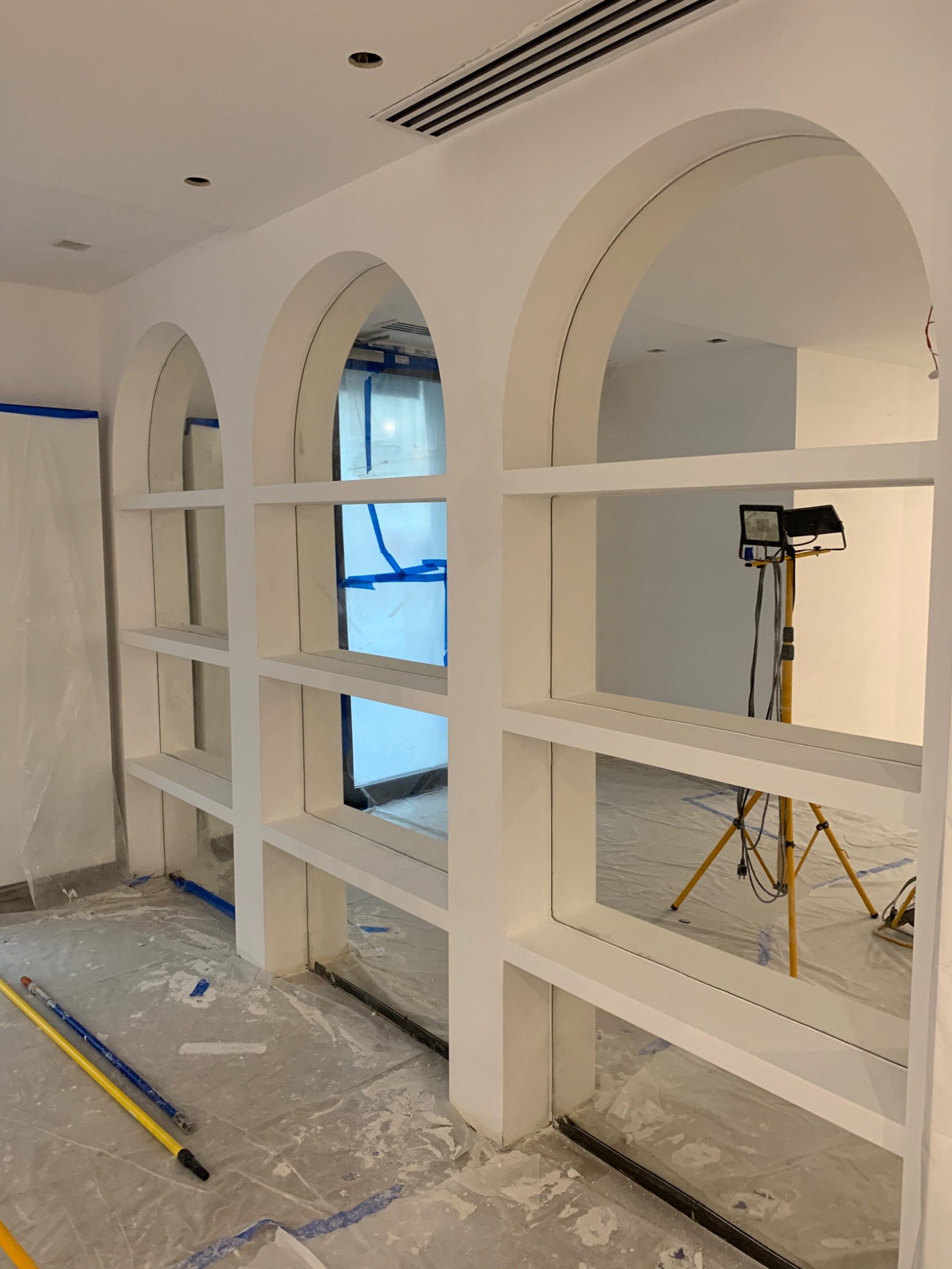 Białe, łukowe półki w trakcie montażu, zabezpieczone folią i taśmą malarską, z widocznym oświetleniem roboczym i narzędziami na podłodze.