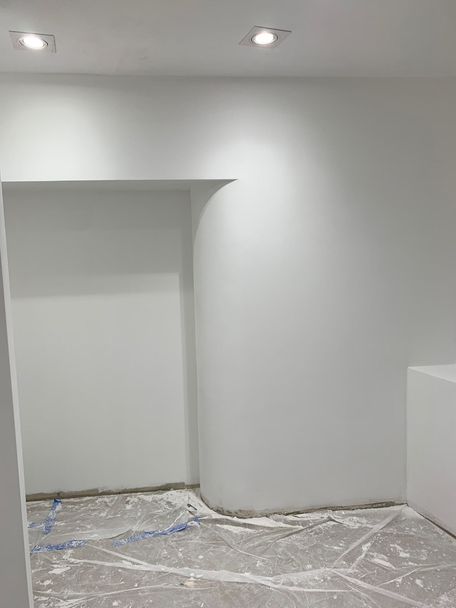 Wnętrze pokoju po świeżym pomalowaniu na biało, z widocznymi plamami farby na folii ochronnej rozłożonej na podłodze i charakterystycznym zaokrąglonym narożnikiem ściany.