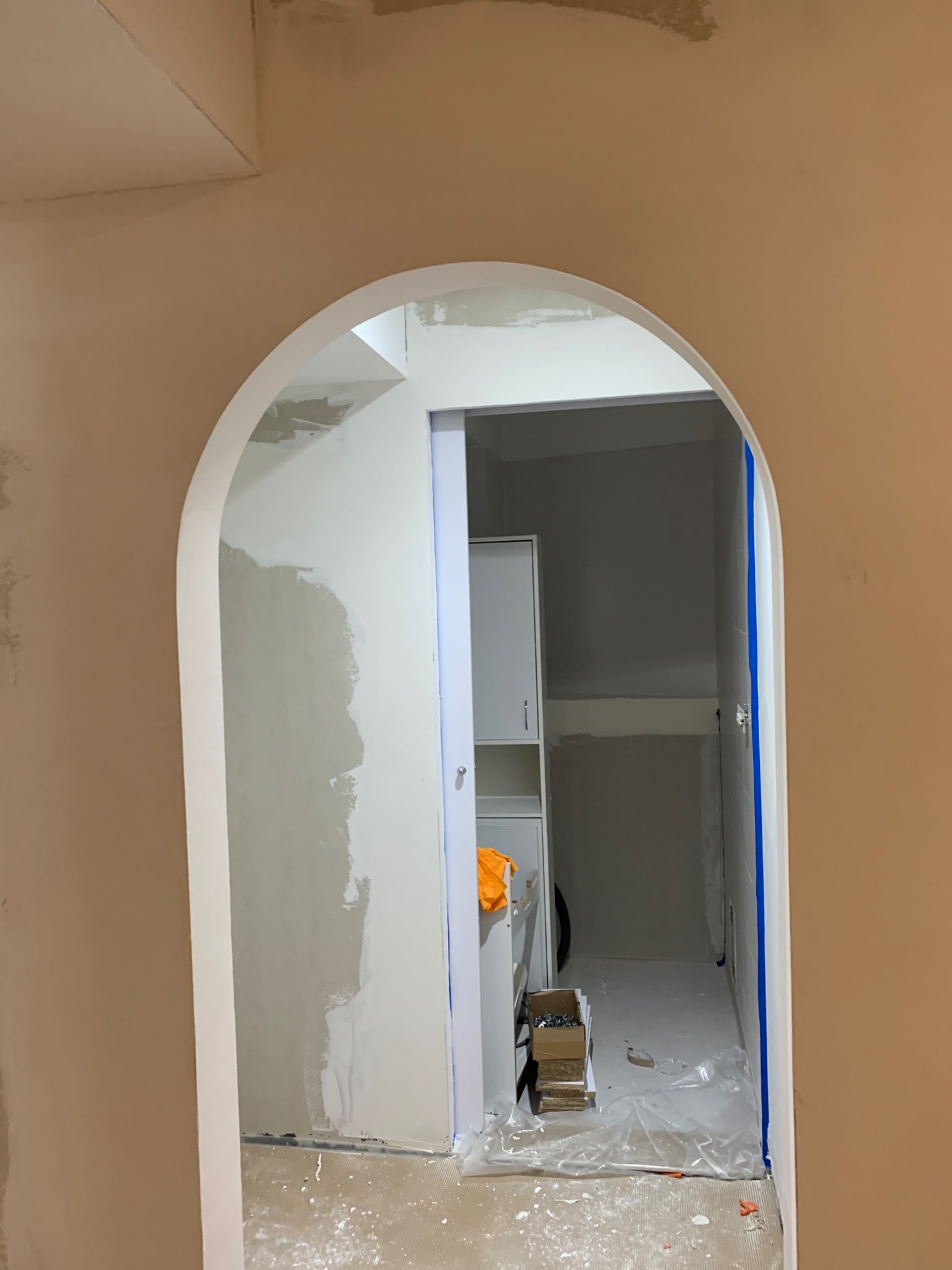 Arched doorway leading to a laundry room under renovation; walls partially plastered, blue painter's tape visible, and a box of screws on the floor.