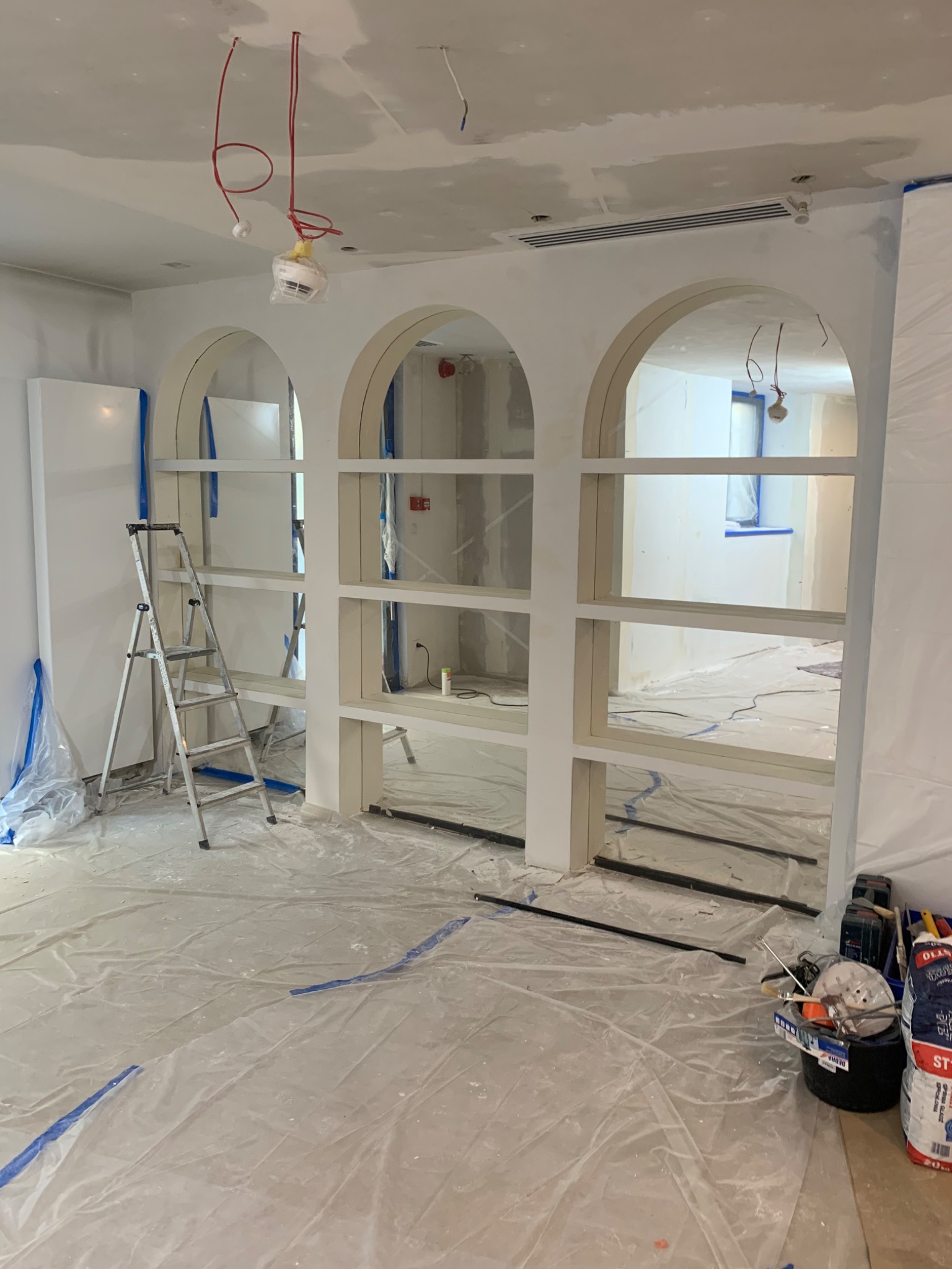 Wnętrze w trakcie prac wykończeniowych: ściany po gładzi, zabudowa z półkami w kształcie łuków, podłoga zabezpieczona folią, drabina i narzędzia malarskie.