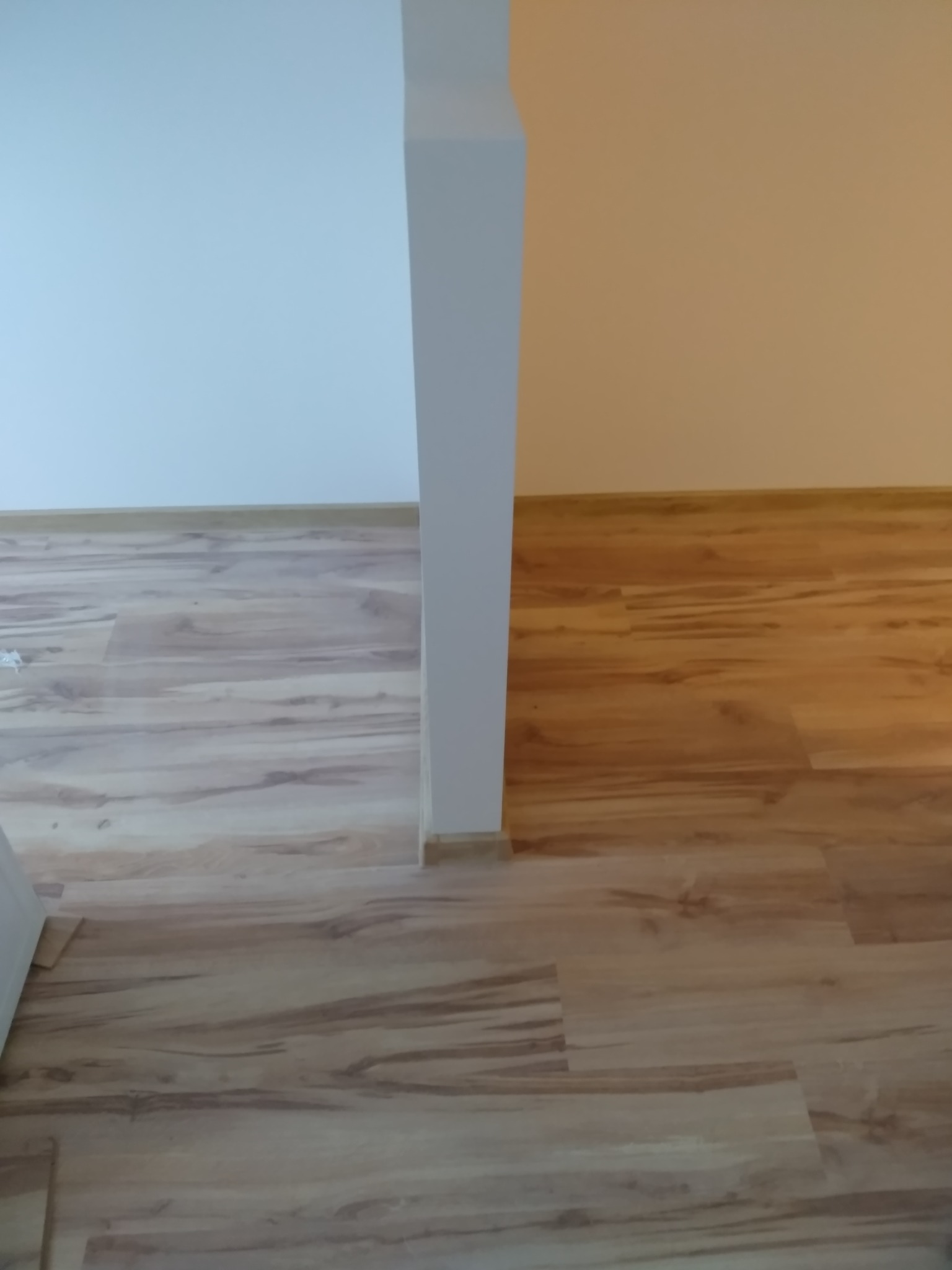 Fragment pokoju z nowo ułożonymi panelami podłogowymi w odcieniach drewna, oddzielonego filarem od ściany pomalowanej na dwa różne kolory: biały i ciepły beż.