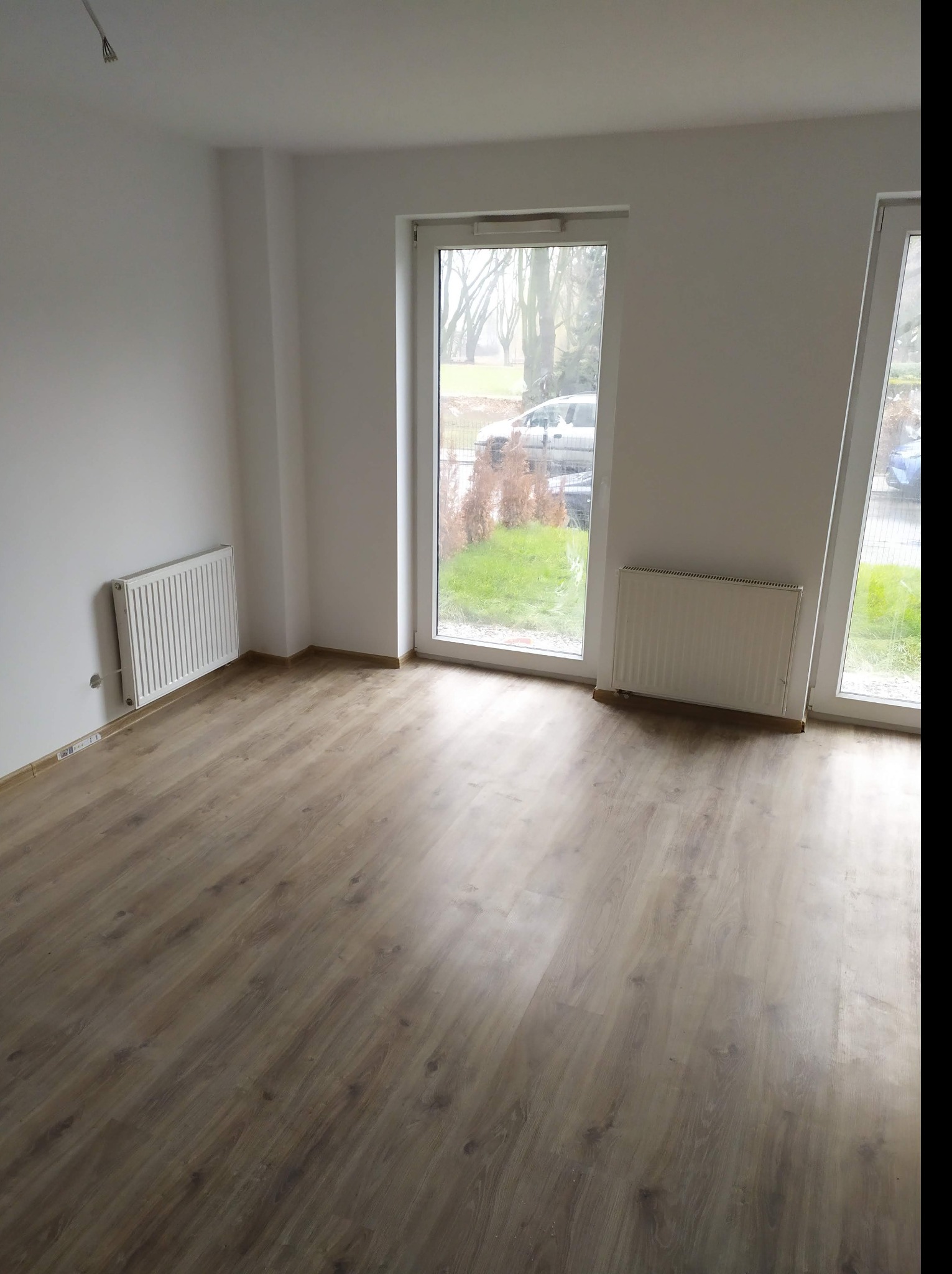 Pusty pokój z panelami podłogowymi w odcieniach brązu, białymi ścianami, grzejnikami i oknami z widokiem na zewnątrz.