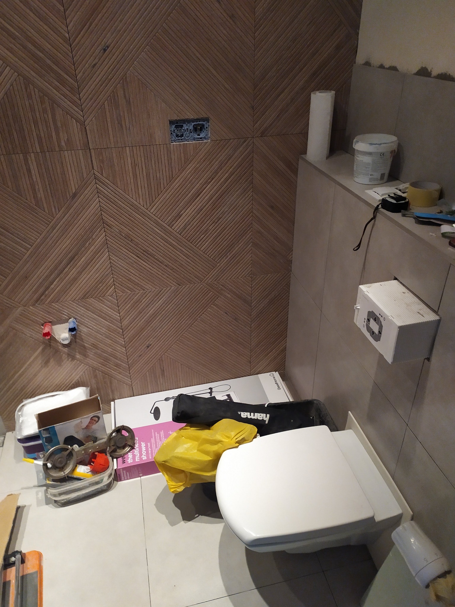 Niedokończona łazienka z drewnopodobną mozaiką na ścianie, widoczne narzędzia, puszka elektryczna bez osprzętu oraz nowa, biała toaleta obok kartonów i folii ochronnej.