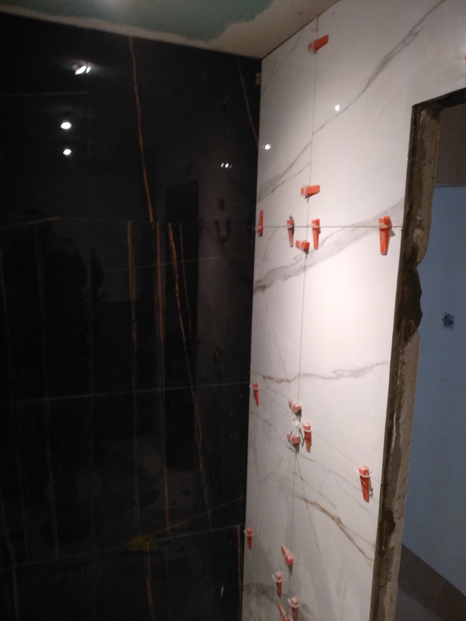 Układanie płytek imitujących marmur na ścianie w łazience, widoczne krzyżyki dystansowe i pionowe sznurki poziomujące.