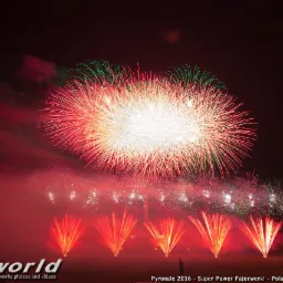 Spektakularny pokaz sztucznych ogni z dominującą czerwienią i zielenią na nocnym niebie, z widocznymi smugami dymu i logo firmy Pyroworld w lewym dolnym rogu.