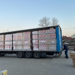 Nordica Transport - Ciężarówka z naczepą wypełniona po brzegi zapakowanymi kartonami, rozładunek w Sokółce, w tle czerwony samochód osobowy z otwartym bagażnikiem i sylwetka pracownika w trakcie pracy.