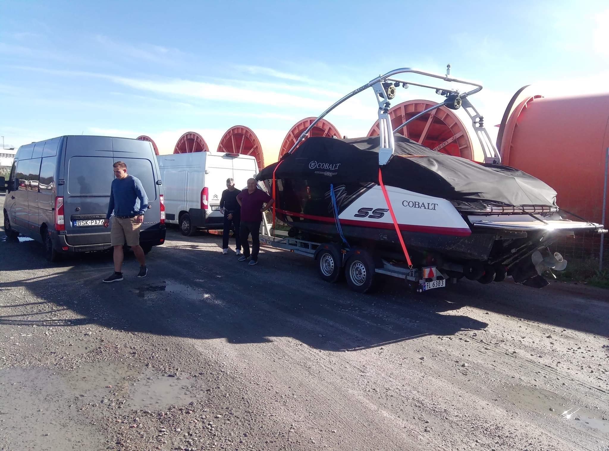 Łódź Cobalt SS na przyczepie, zabezpieczona czarną plandeką i czerwonymi pasami, transportowana przez szarą furgonetkę Renault Master, z widocznymi osobami i bębnami kablowymi w tle.