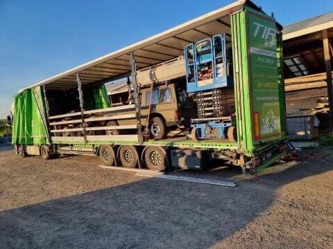 Otwarta naczepa ciężarówki z zieloną plandeką, przewożąca samochód terenowy, podnośnik koszowy i inne ładunki.