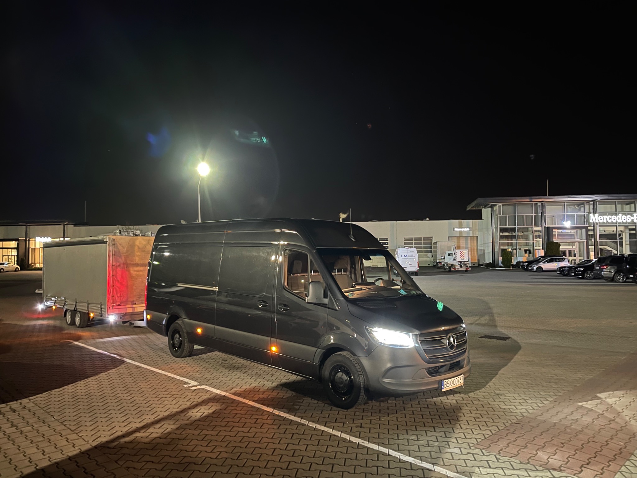 Szary Mercedes Sprinter z przyczepą towarową zaparkowany na placu w nocy, oświetlony światłami ulicznymi. Widoczne budynki w tle.