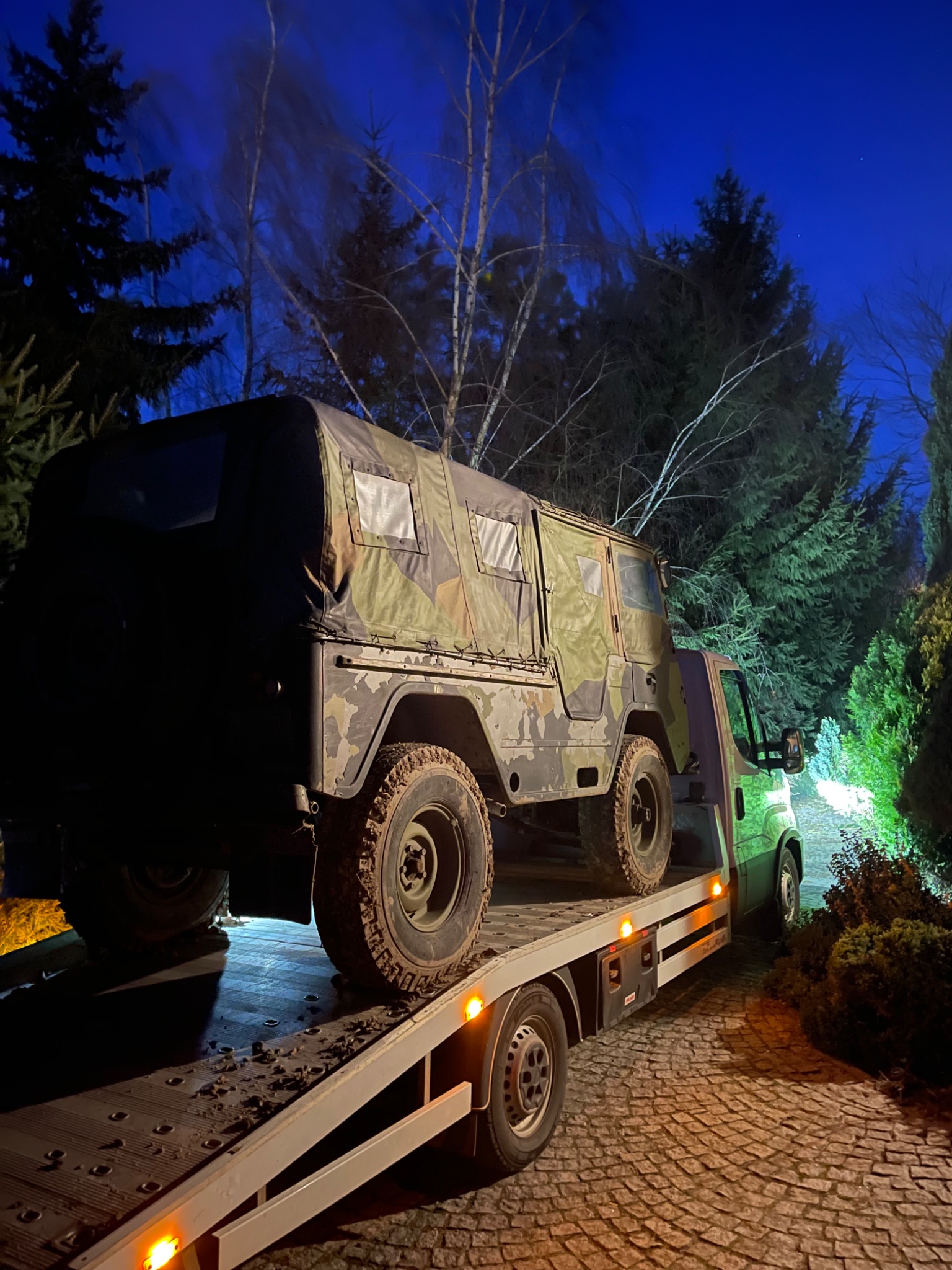 Ciężarówka transportowa z wojskowym pojazdem terenowym w kamuflażu na platformie, nocne ujęcie z oświetleniem, widoczne błoto na oponach.